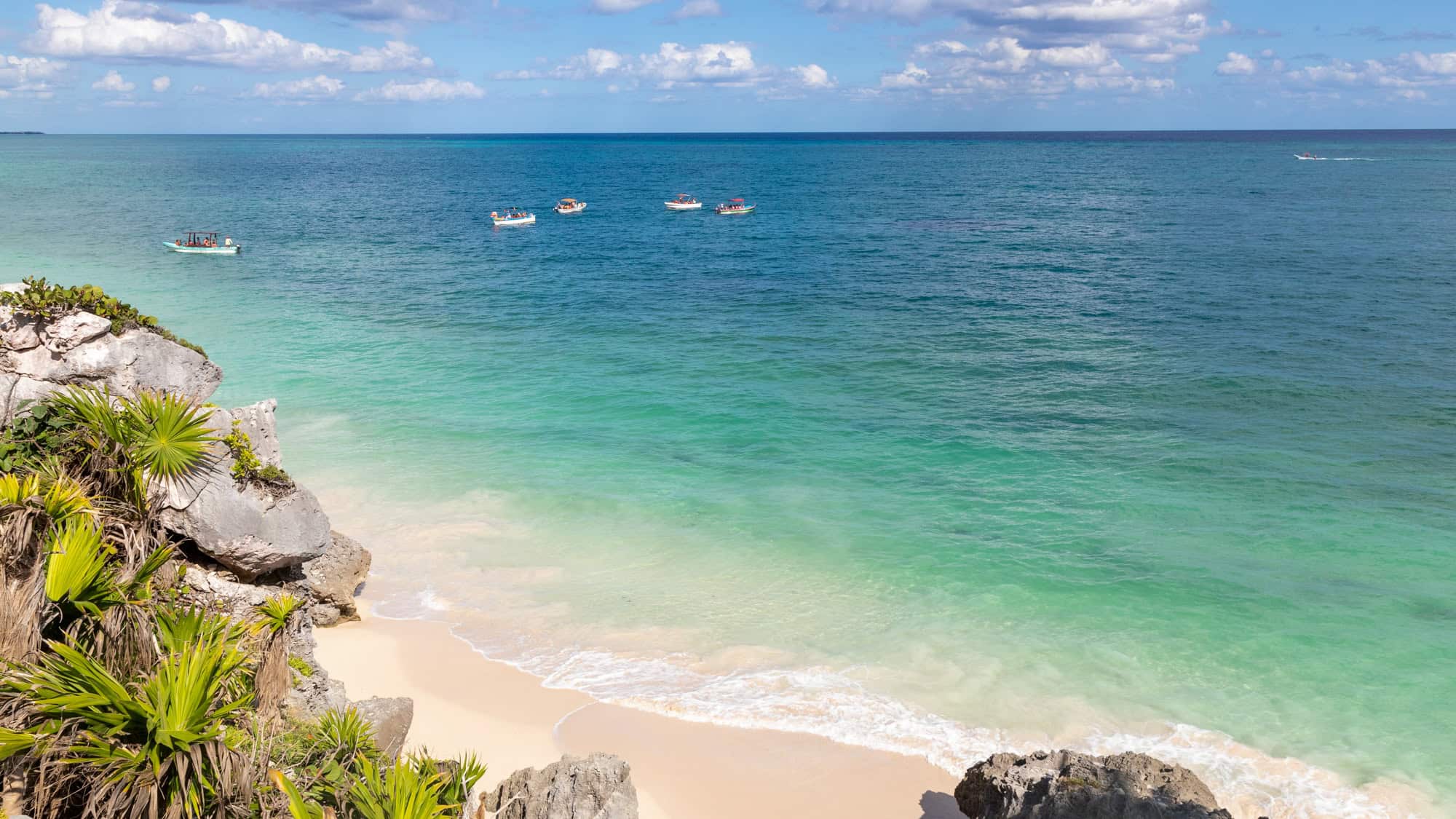 A sandy beach with turquoise waves laps against rocky cliffs and palm trees, while small boats float offshore.