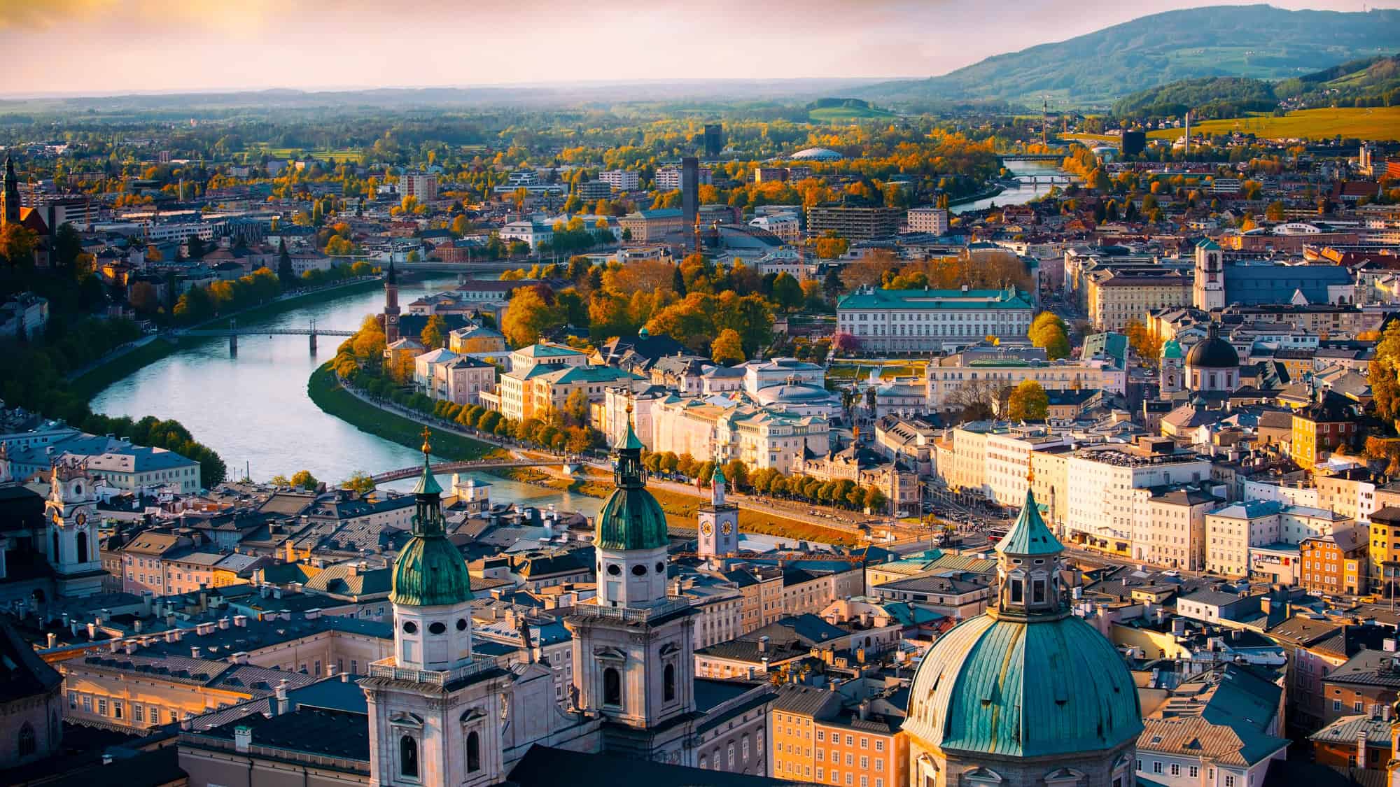 The Salzach River winds through Salzburg, Austria, with church domes and historic buildings glowing in golden evening light.