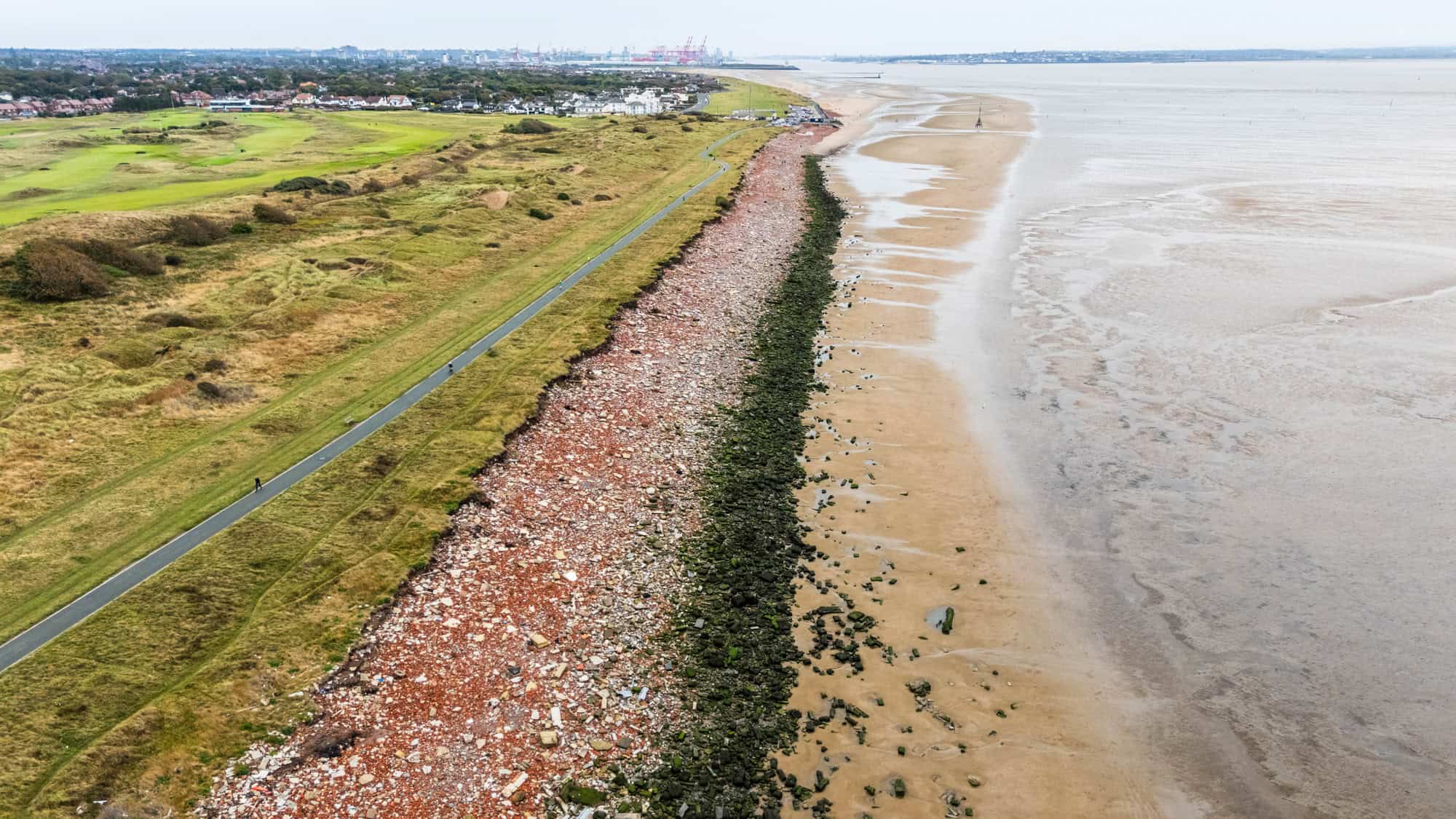 A long coastal path runs parallel to a beach with a stretch of red rubble and mossy rocks, separating grassy fields from wide tidal flats.
