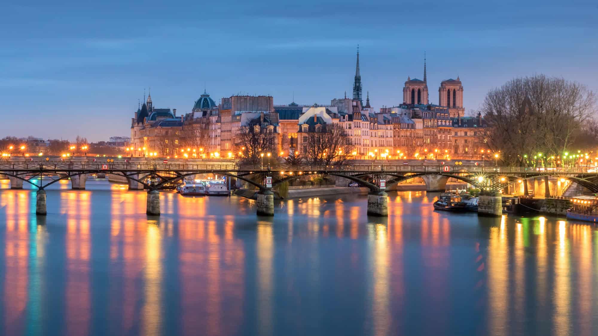 Lights shimmer on the Seine River as bridges lead toward the illuminated Notre-Dame Cathedral at twilight.