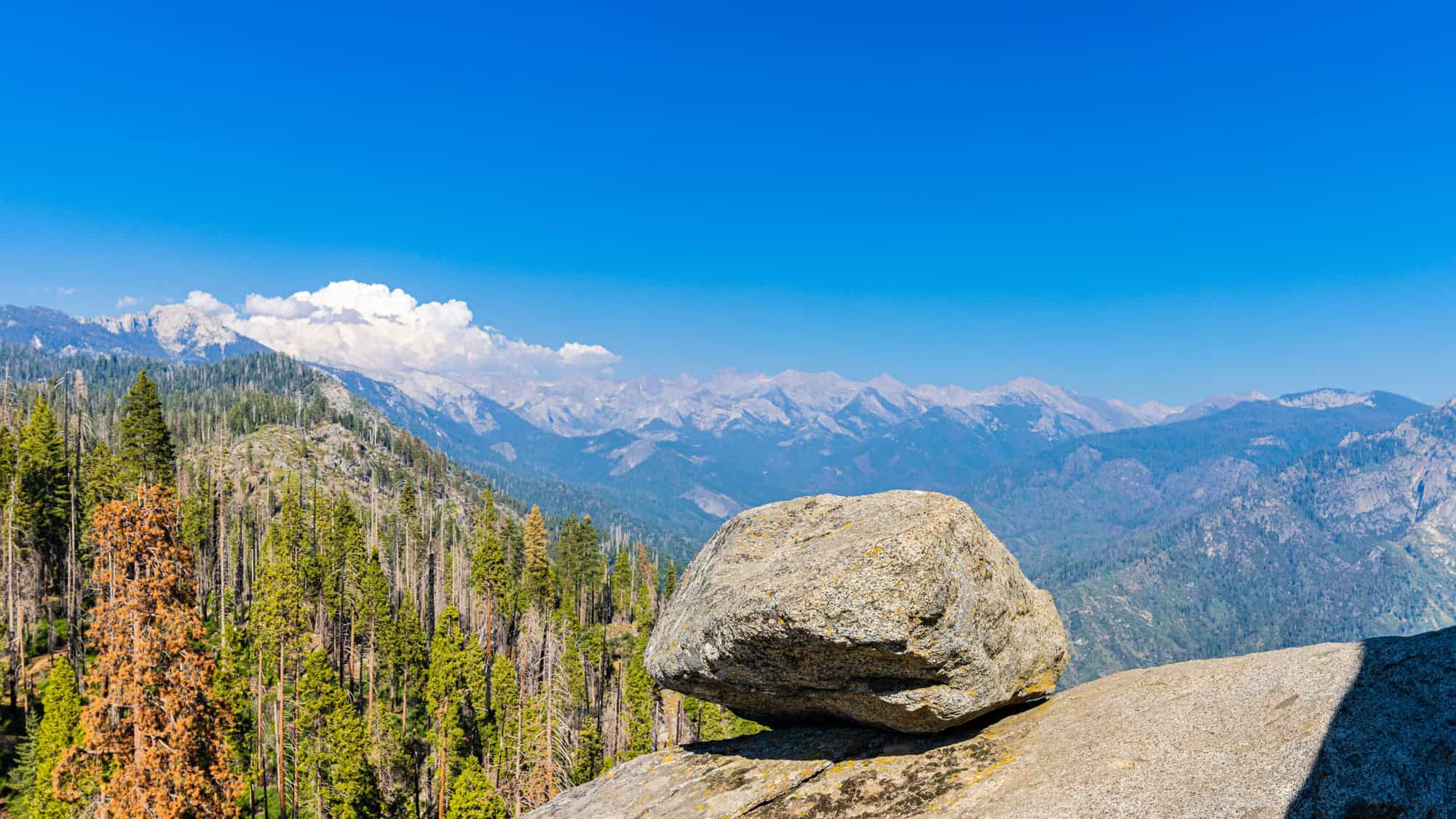 A large boulder rests on a rocky ledge overlooking forested mountains under a vivid blue sky.