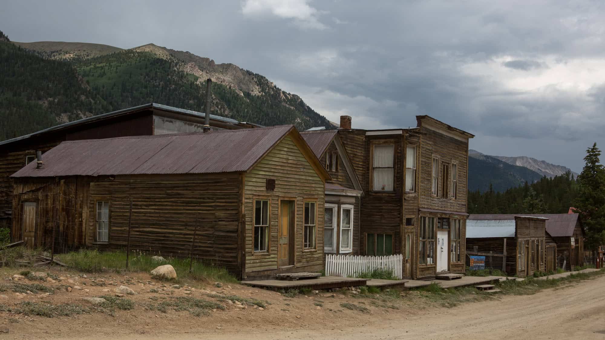 Weathered wooden buildings with false fronts and metal roofs line a dirt street in the former mining town of St. Elmo, Colorado, beneath stormy mountain skies.