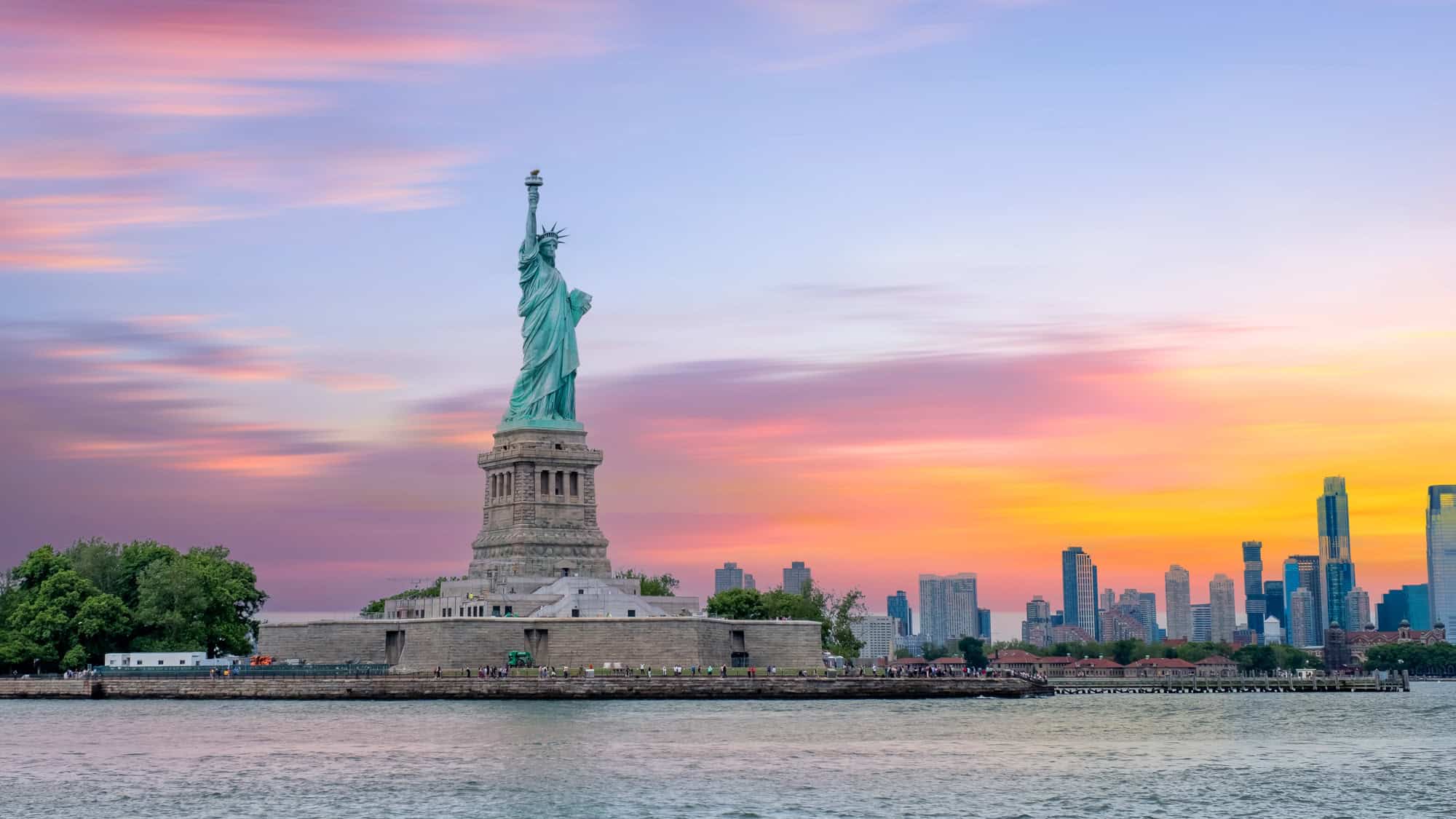 The Statue of Liberty towers over the harbor at sunset with New York City’s skyline in the background.