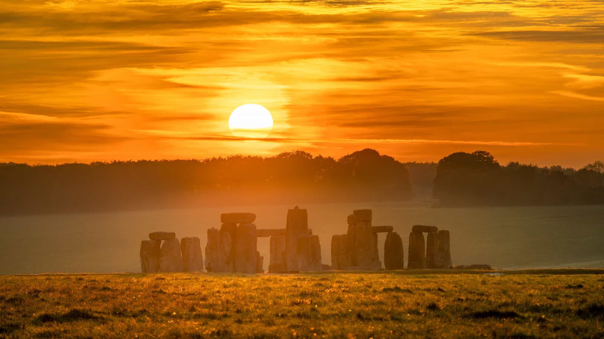 The ancient stone circle of Stonehenge is silhouetted against a brilliant orange sunset sky.