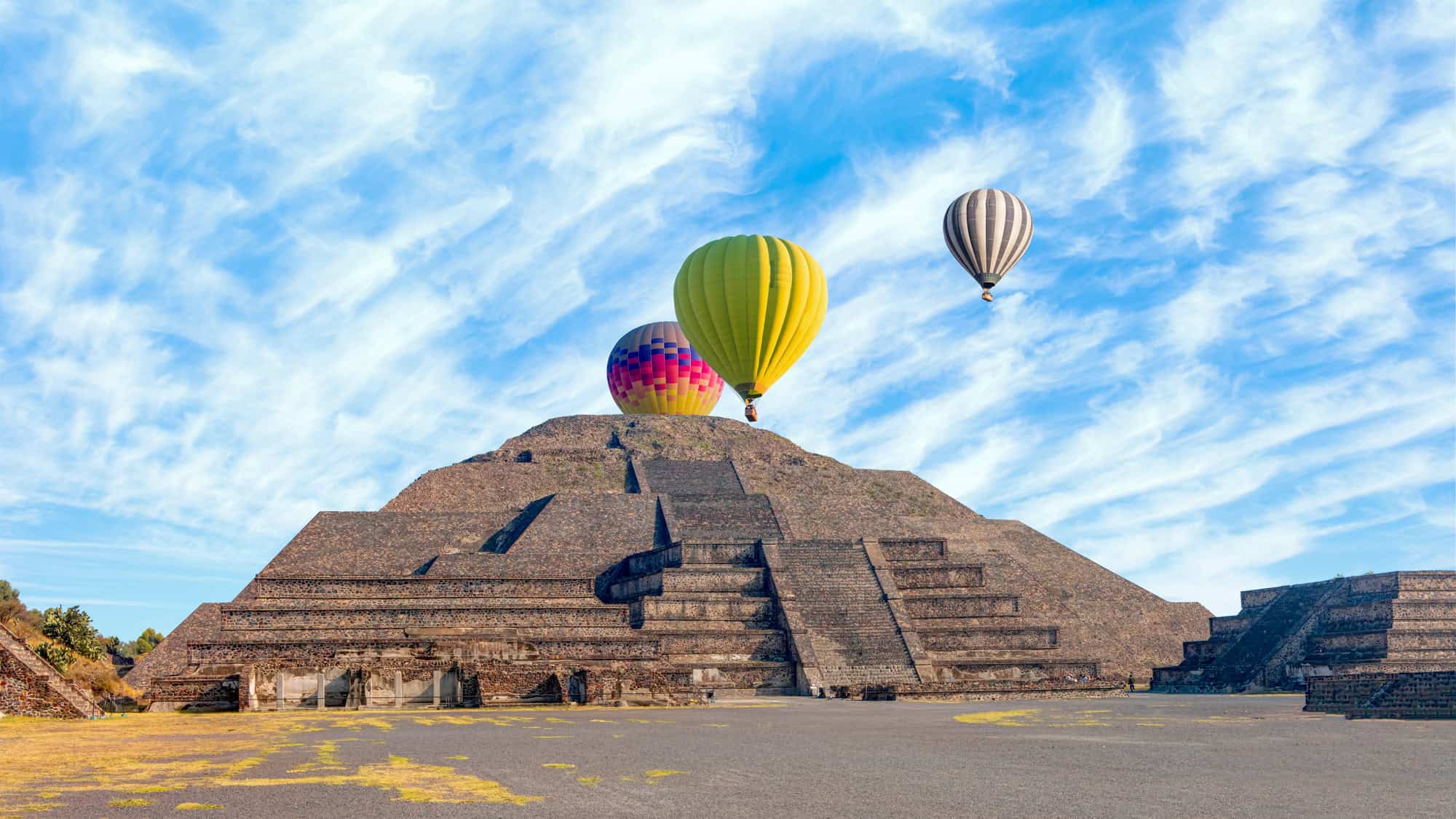 Hot air balloons float above the Pyramid of the Sun at Teotihuacan under a sky streaked with wispy clouds.