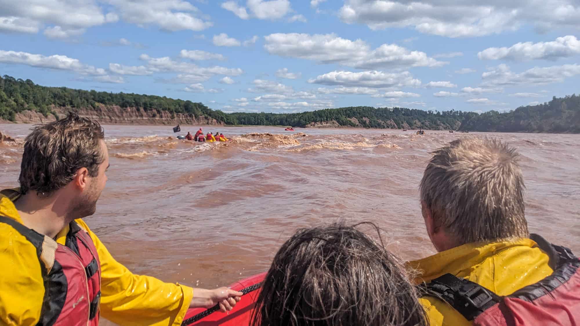 Rafters in yellow gear navigate through fast-moving, muddy river rapids under a bright sky.