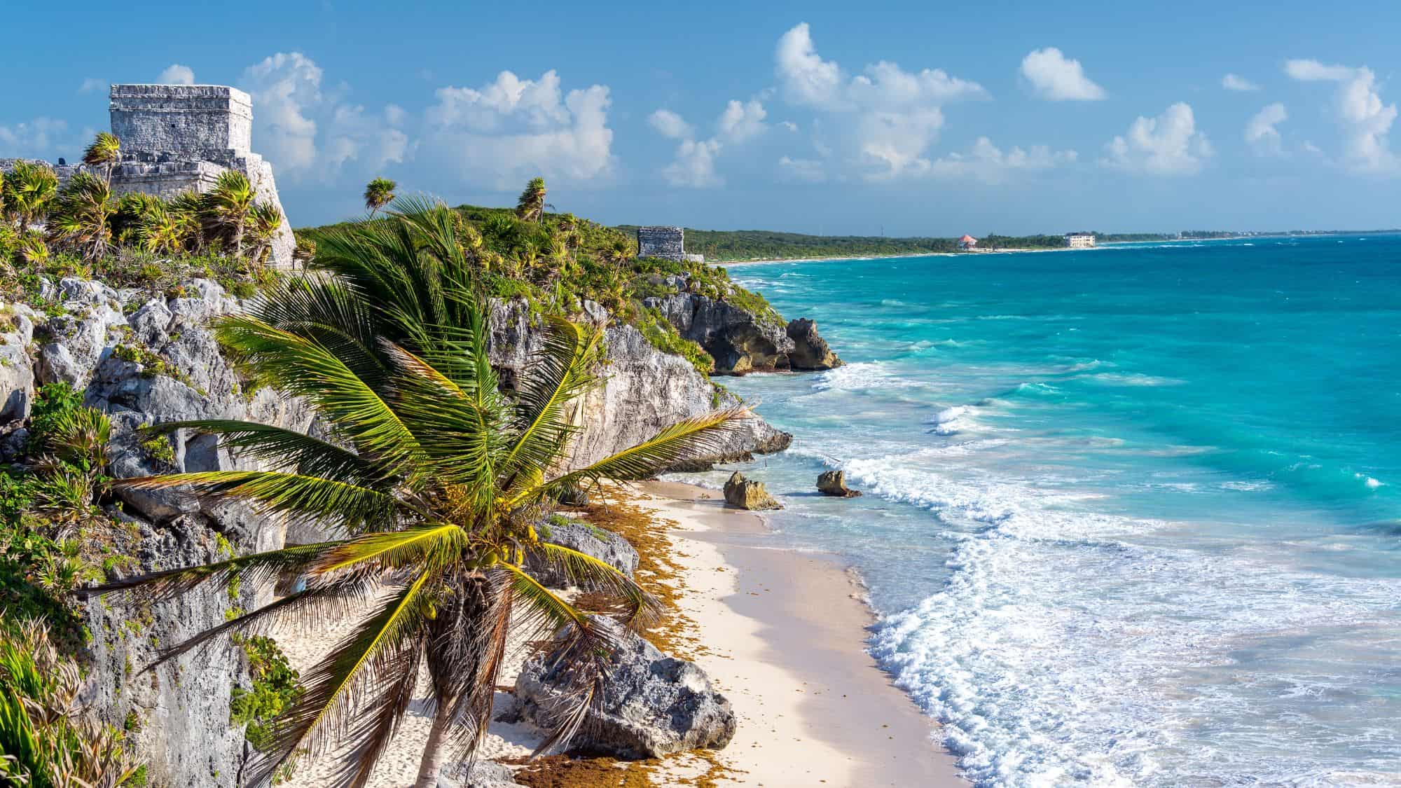 Ancient Mayan stone temples sit atop cliffs overlooking turquoise waves and palm trees along the Caribbean coast.