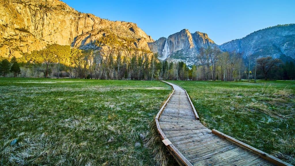 A wooden boardwalk stretches across a green meadow toward towering granite cliffs and a tall waterfall.
