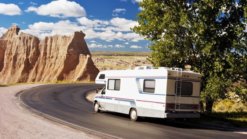 A white RV winds along a curving road through the rugged, eroded cliffs of Badlands National Park, with expansive views stretching into the distance.