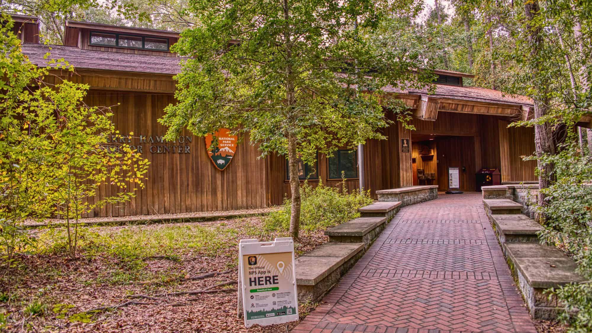 A wooden building labeled "Kim Hampton Visitor Center" is nestled among trees, with a brick walkway leading to the entrance and a sign promoting the official NPS app.