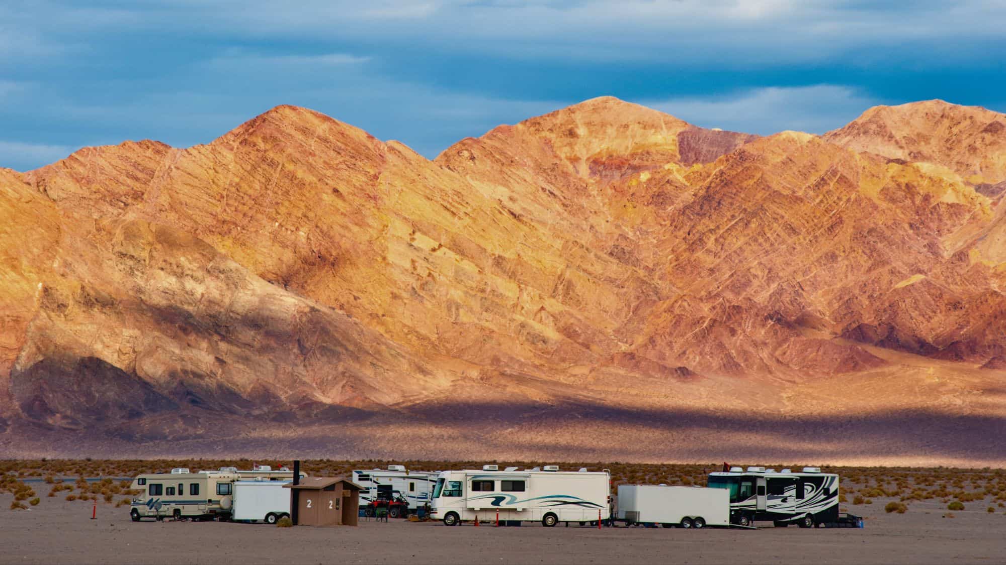 A cluster of RVs parks in the wide open desert beneath strikingly layered and colorful rocky mountains under a moody sky.
