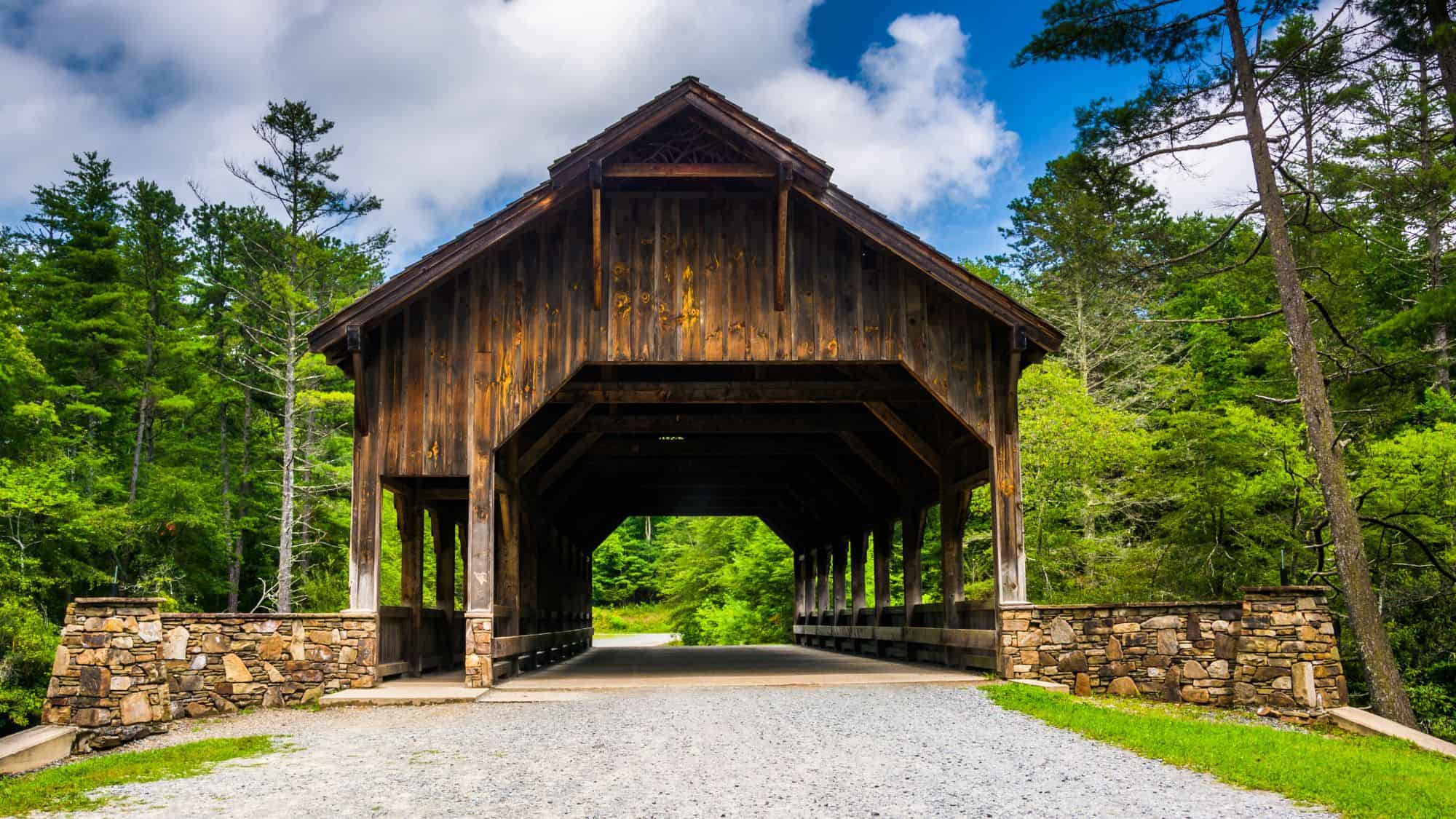 A rustic wooden covered bridge with stone supports stands under a partly cloudy sky, framed by lush green trees on both sides.