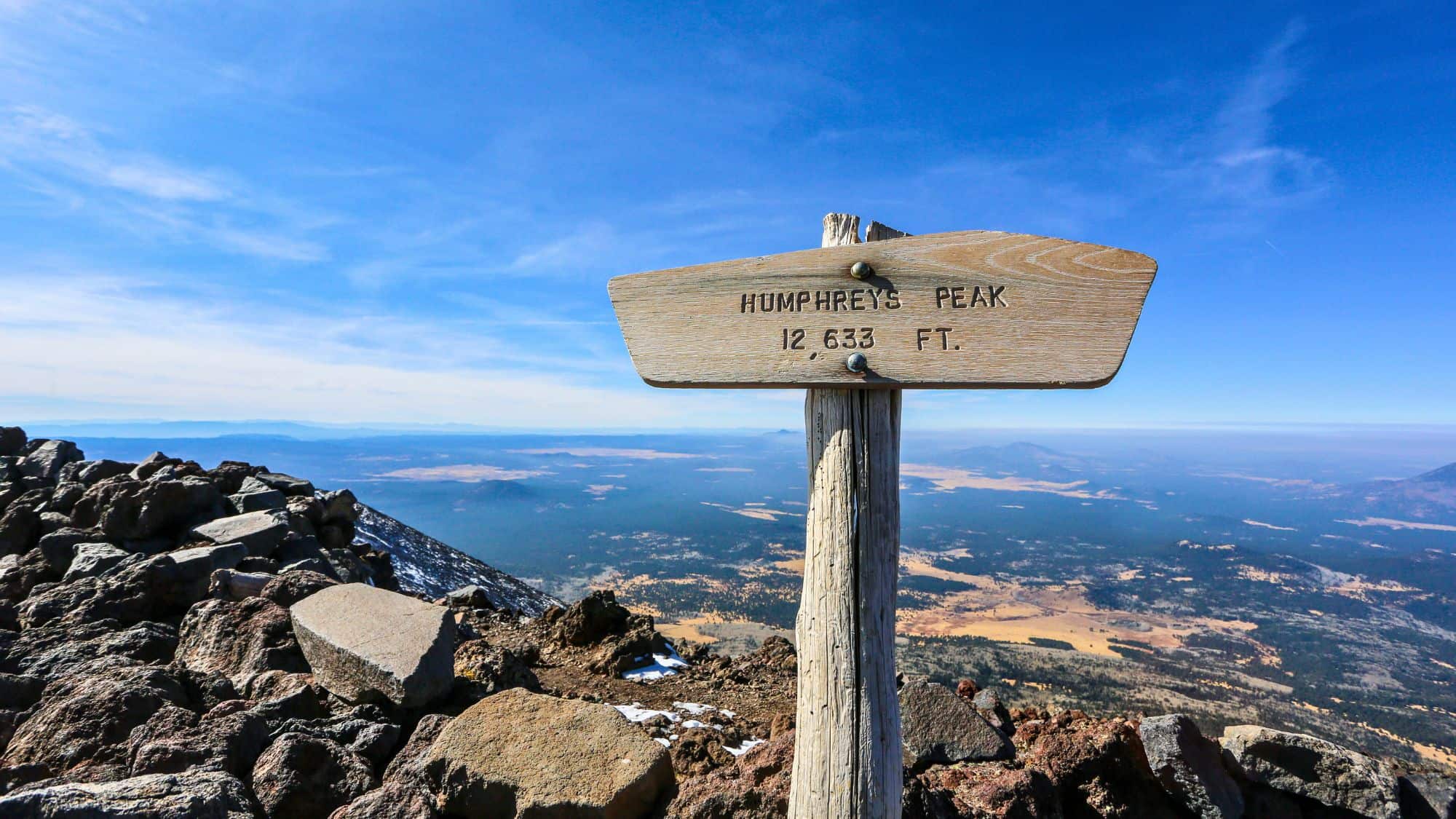 A weathered wooden sign marks the summit of Arizona’s highest peak at 12,633 feet, with expansive views of the surrounding forested landscape and distant mountains below.