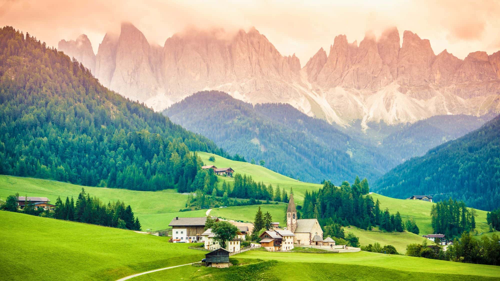 A charming mountain village with a church steeple sits nestled in green hills, backed by forested slopes and the jagged Dolomites glowing in soft morning light.