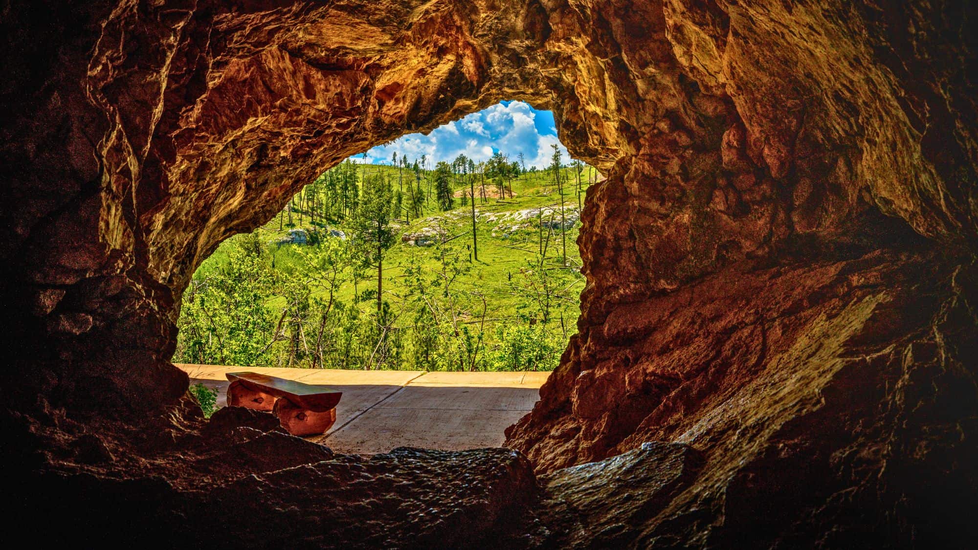 Looking out from the mouth of a rocky cave, a lush green hillside dotted with trees and rocks is framed beneath a bright blue sky with puffy clouds.