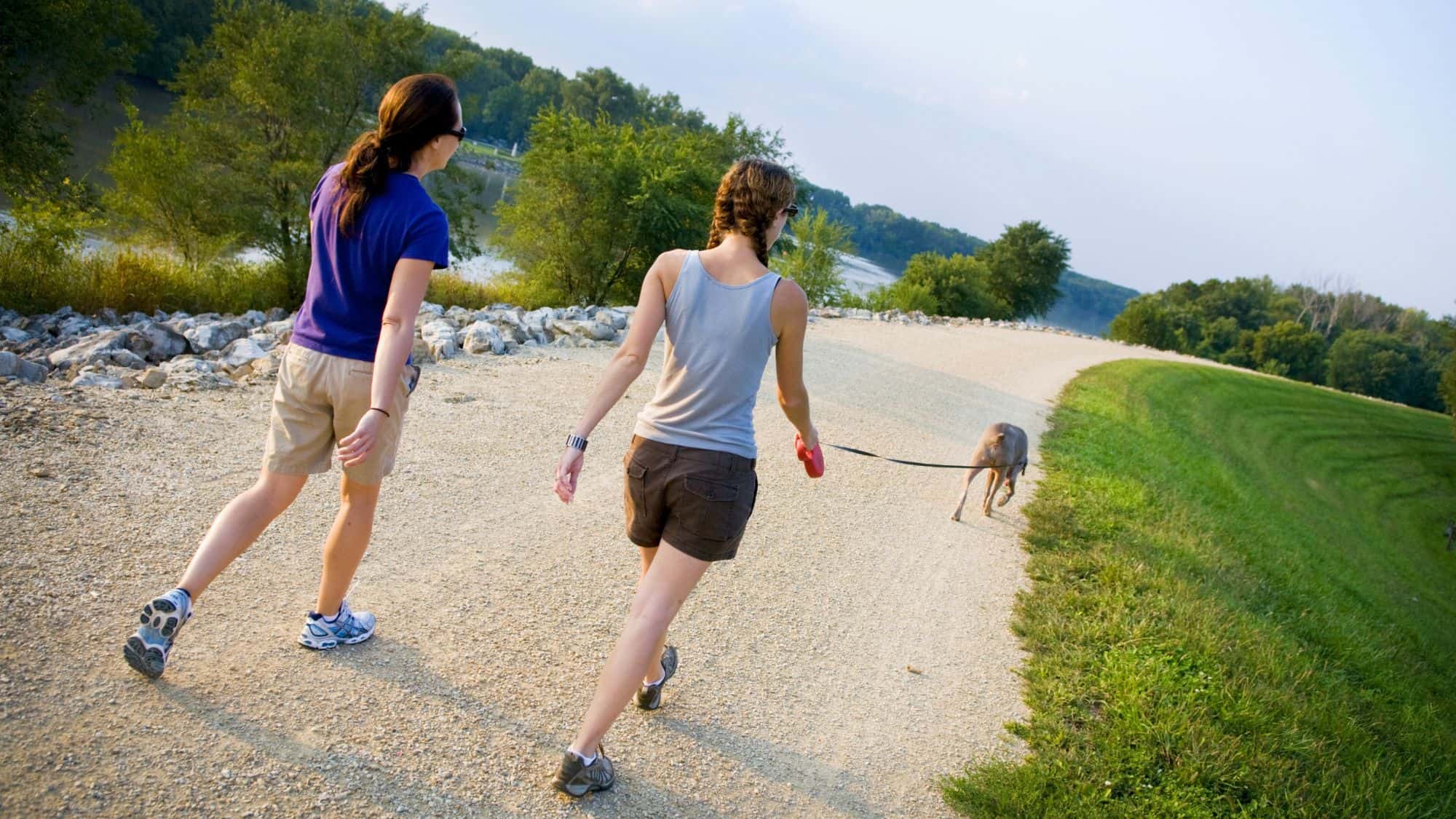 Two women walk a dog along a curving gravel path by a river, surrounded by green grass, rocks, and trees under a clear sky.