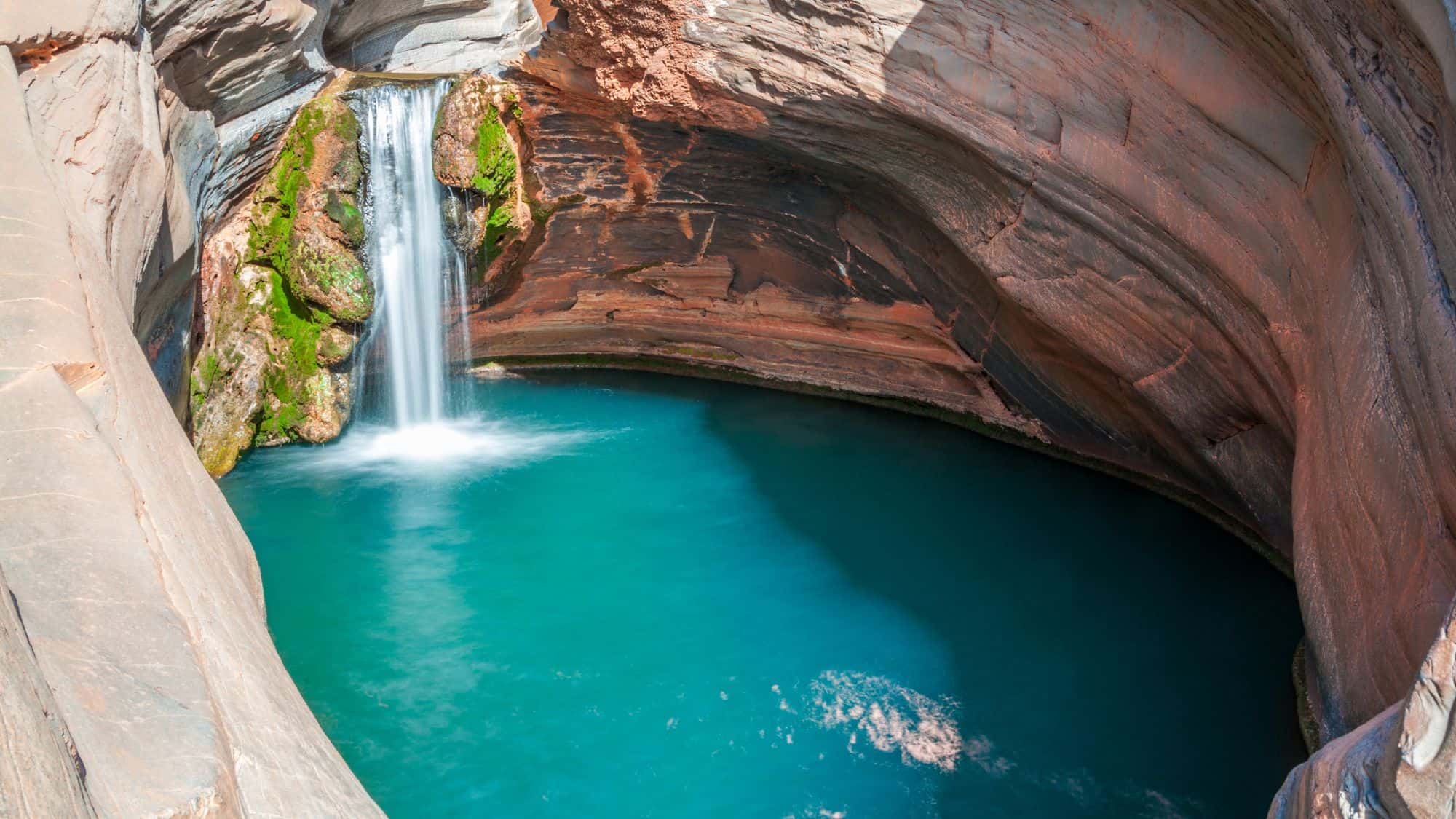 A small waterfall cascades into a vibrant turquoise pool enclosed by smooth, curved canyon walls streaked with red and gray rock.