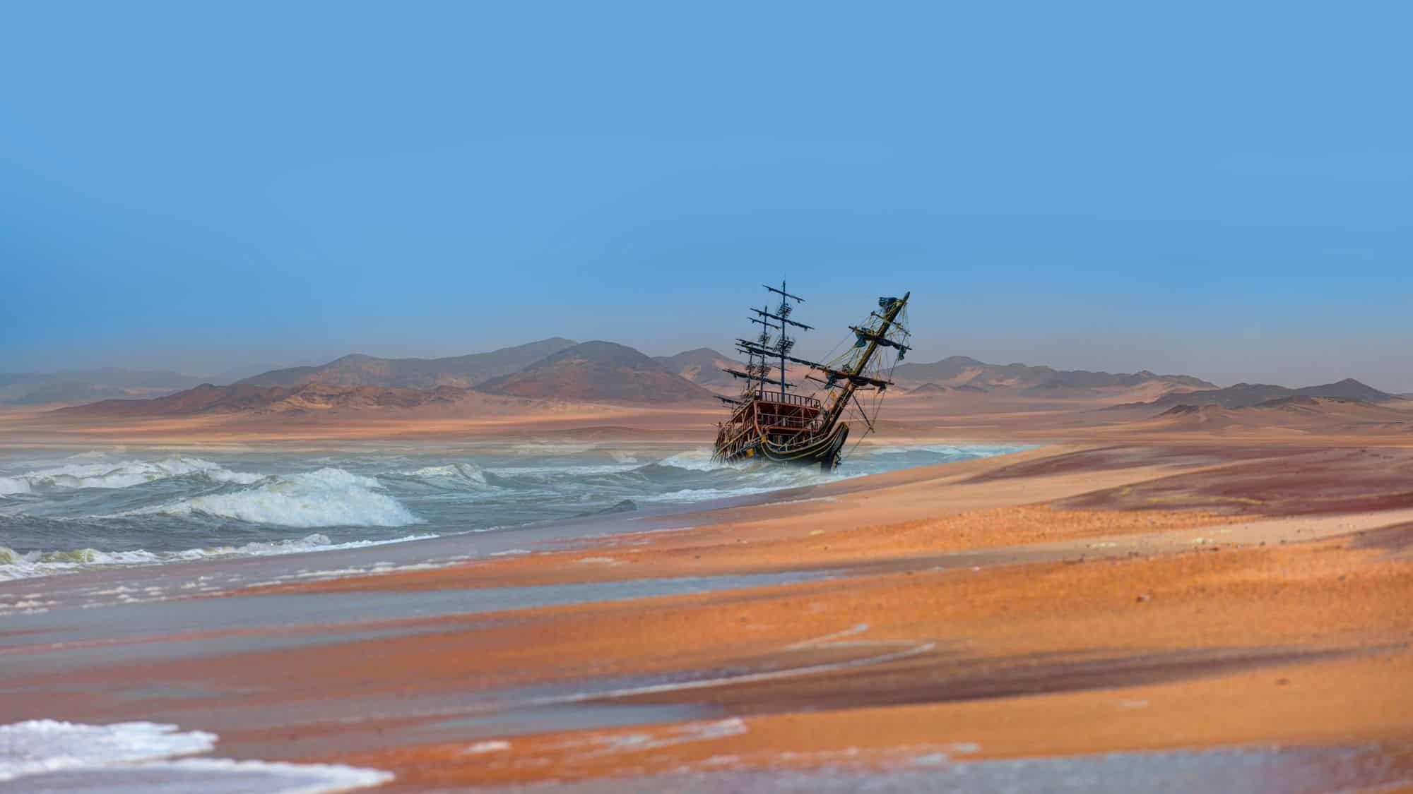 An old, weathered shipwreck leans in the surf along Namibia’s Skeleton Coast, with rust-colored sand dunes and hazy desert mountains in the distance.