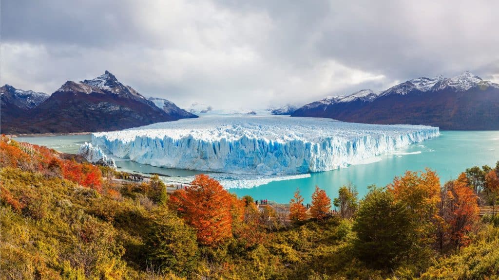 Tourists gather on wooden walkways overlooking the massive, jagged ice face of the Perito Moreno Glacier, surrounded by fiery autumn foliage and snow-dusted mountains.