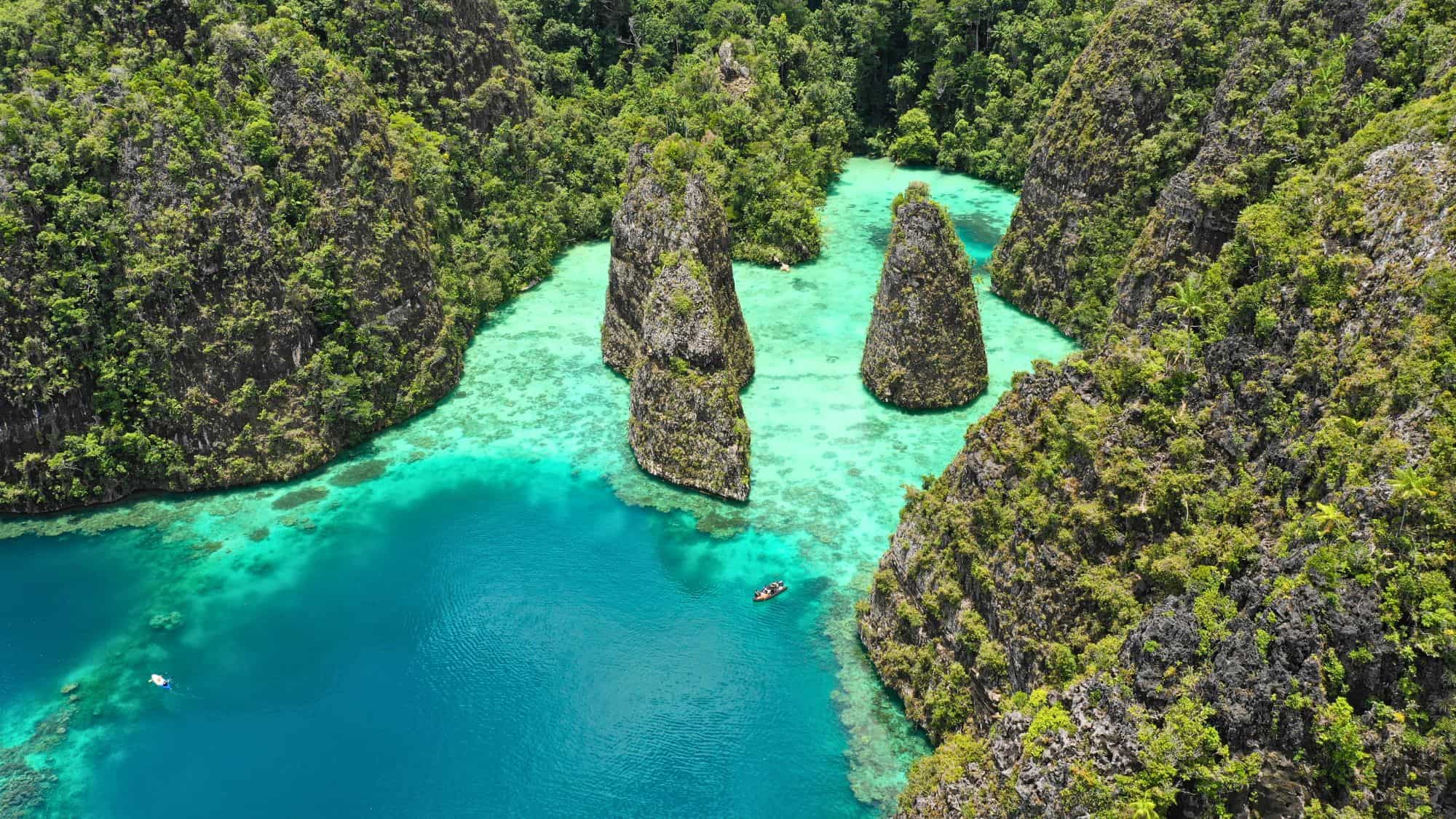 Towering limestone karst formations rise from a glowing turquoise lagoon surrounded by dense jungle, with small boats drifting below.