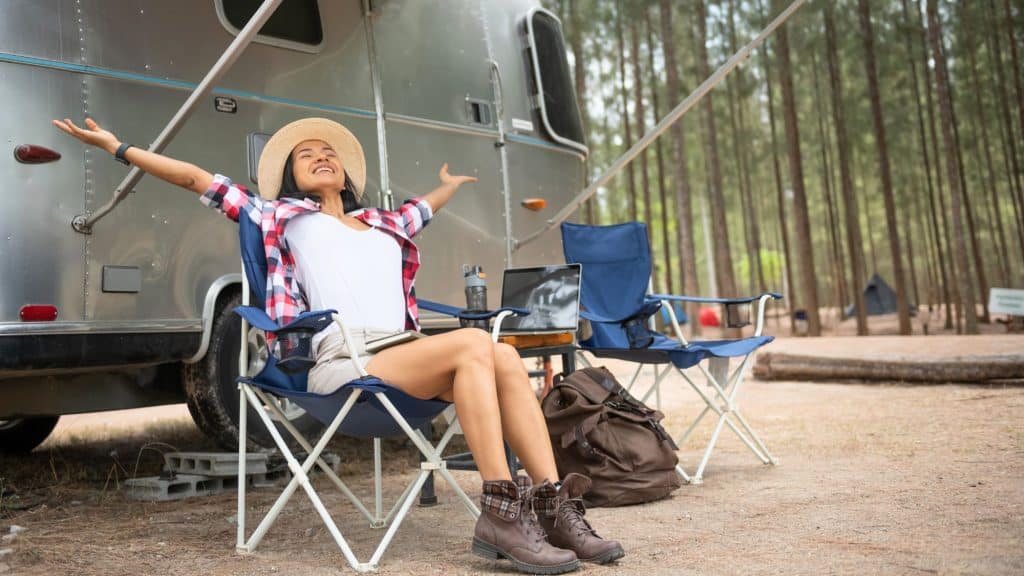 A woman in hiking boots and a straw hat relaxes in a camp chair outside an Airstream trailer, stretching her arms with joy in a peaceful pine forest.