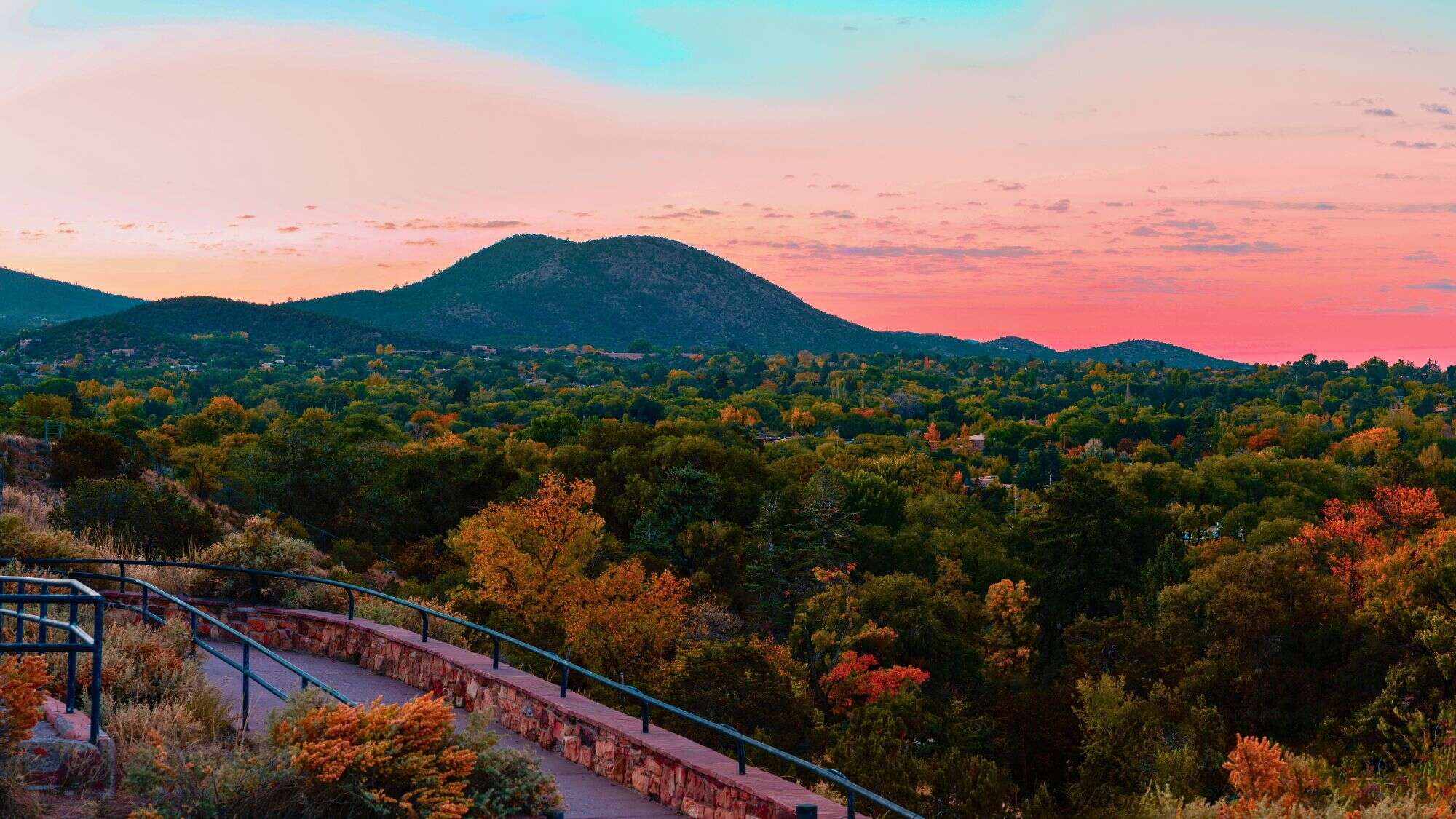 A paved trail with metal railings curves through autumn-colored trees toward distant rolling hills under a vibrant pink and blue sunset sky.