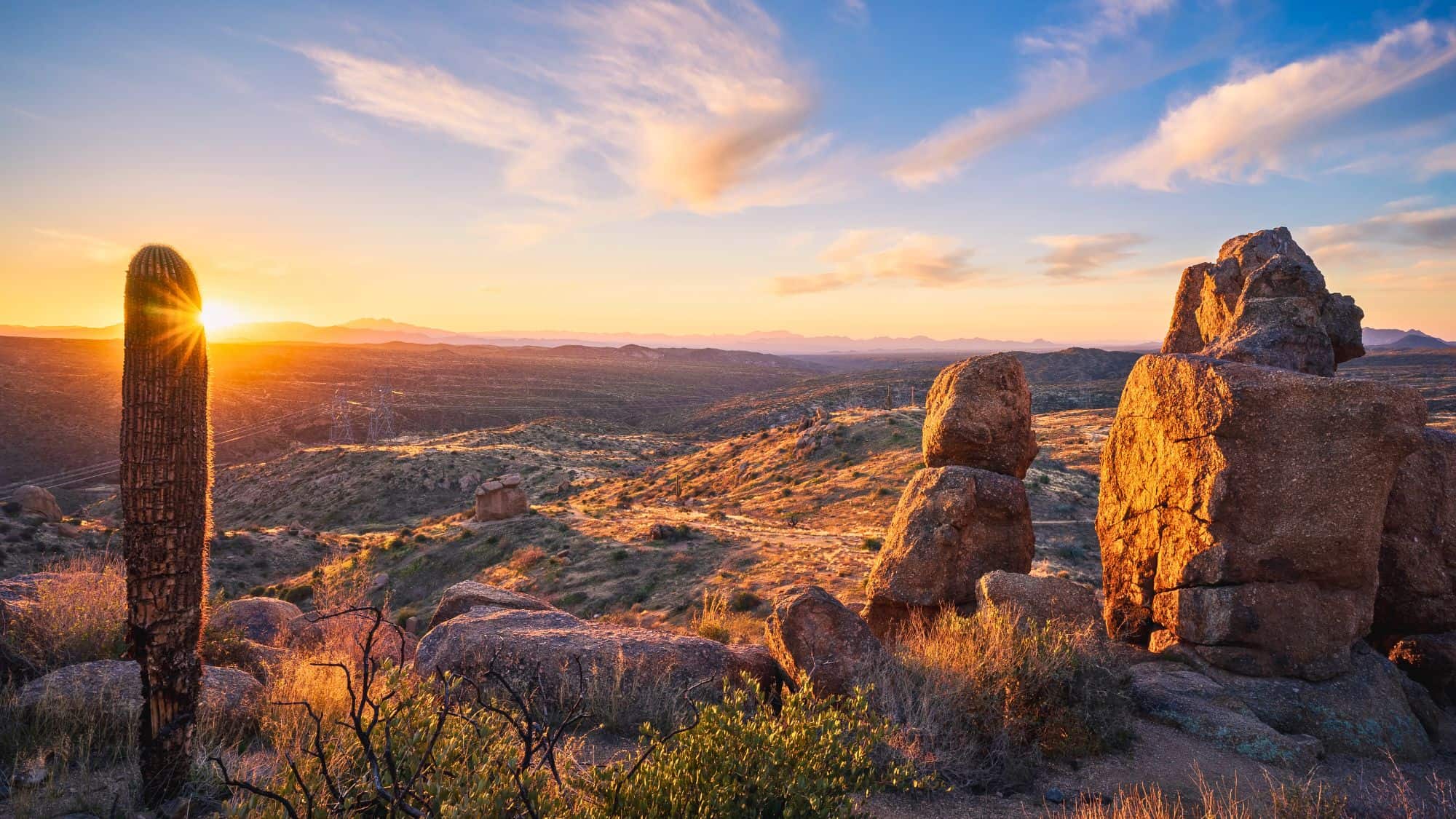 Golden sunlight breaks through a tall saguaro cactus in a rocky, hilly desert at sunset, casting warm tones across the vast Sonoran landscape.