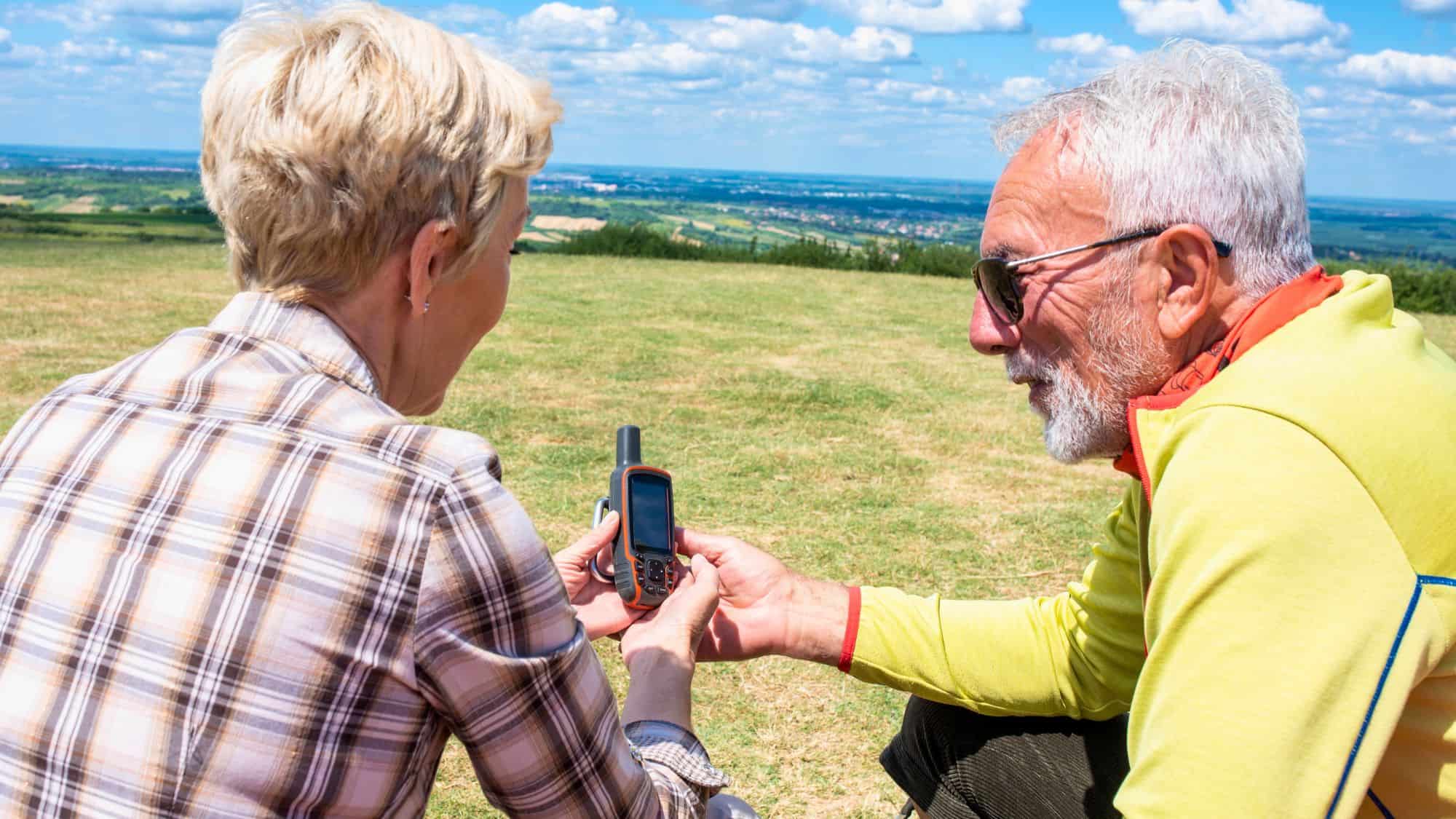 An older couple sits on a grassy hilltop looking at a handheld GPS device with a scenic landscape in the background.