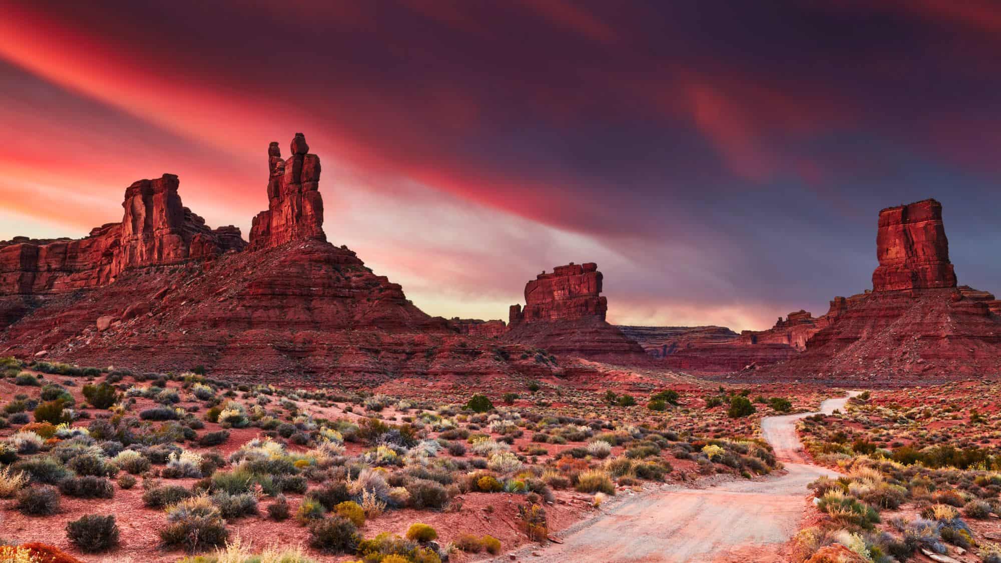 Red sandstone formations rise from the desert floor in the Valley of the Gods, Utah, under a dramatic sky streaked with glowing pink and purple clouds.