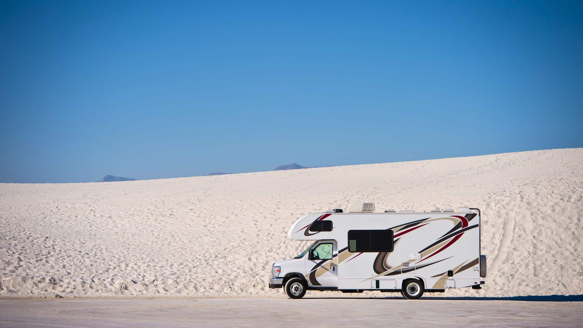 A white RV is parked against a vast backdrop of soft, rolling white sand dunes under a cloudless blue sky, creating a scene of minimalist desert travel.