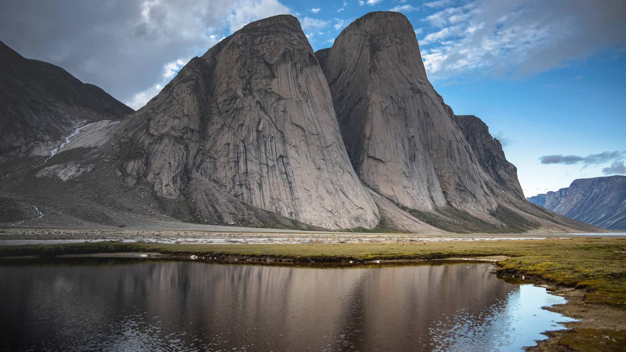 Massive, sheer rock faces known as the Twin Peaks rise dramatically from a glacier-fed valley, reflected in a calm pool under a partly cloudy sky.