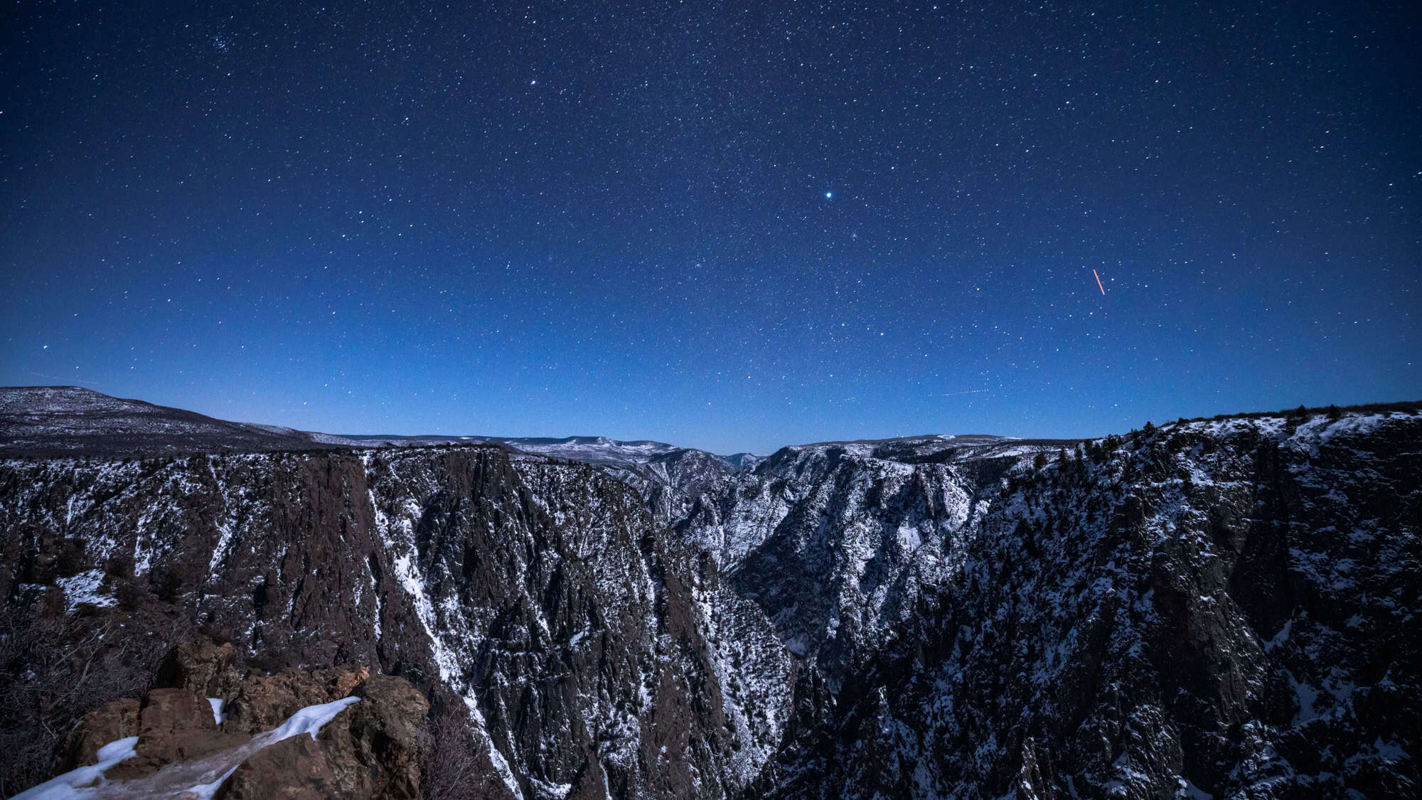 A dramatic, snow-dusted canyon stretches between steep cliffs beneath a crystal-clear night sky filled with stars and a faint meteor streak.