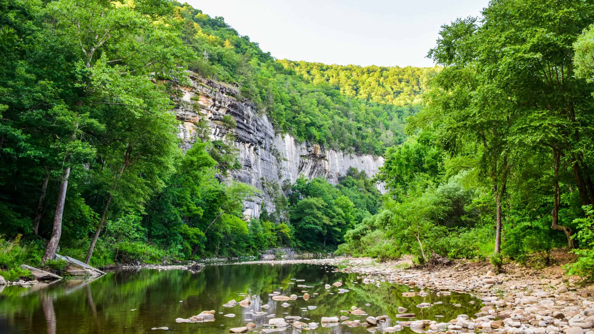 A calm river winds through a lush green forest beneath a towering rock bluff, with smooth stones lining the shallow riverbank.