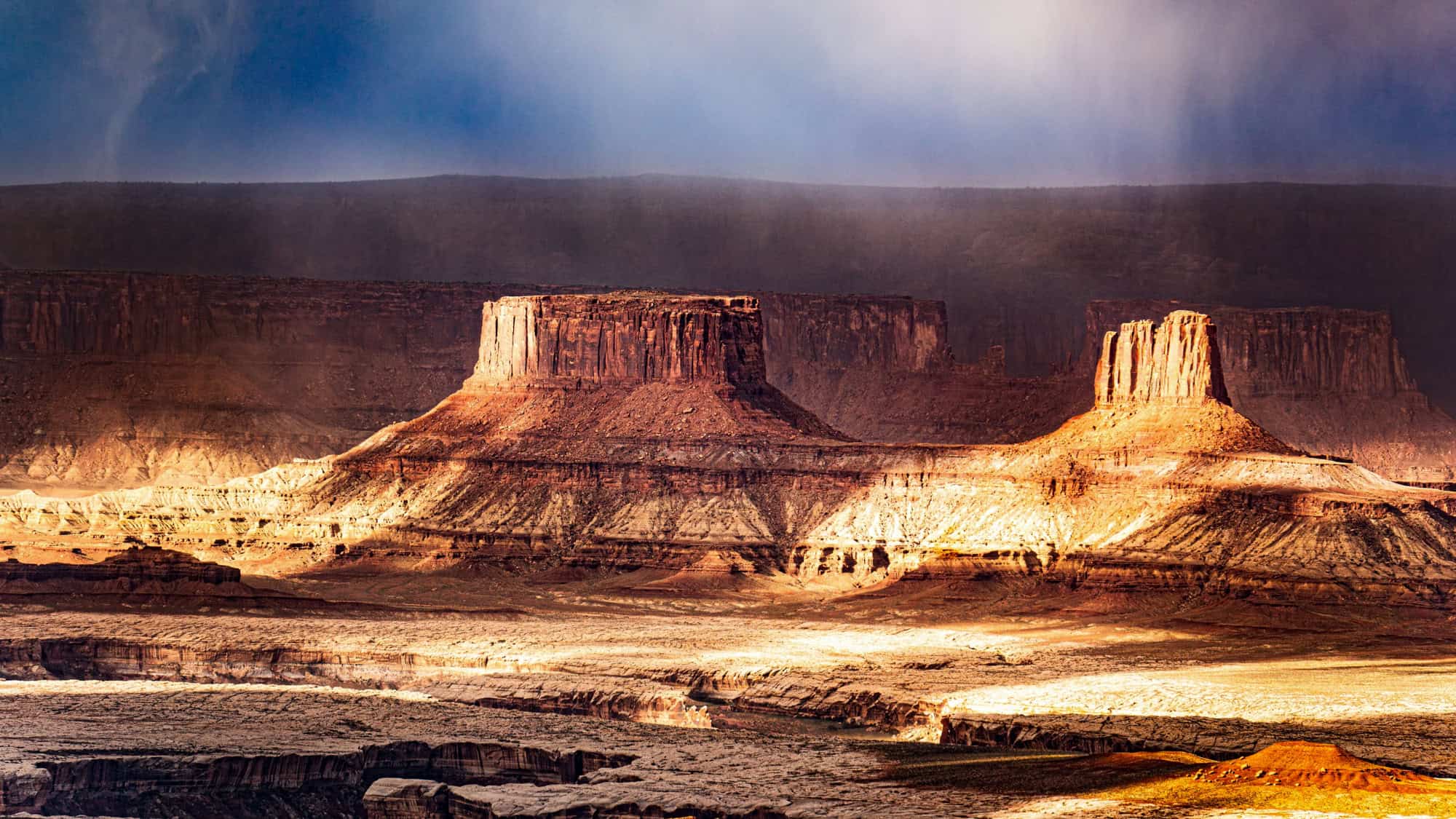 Two flat-topped mesas glow under filtered sunlight in Canyonlands National Park, with dramatic shadows and storm clouds adding depth to the layered desert landscape.