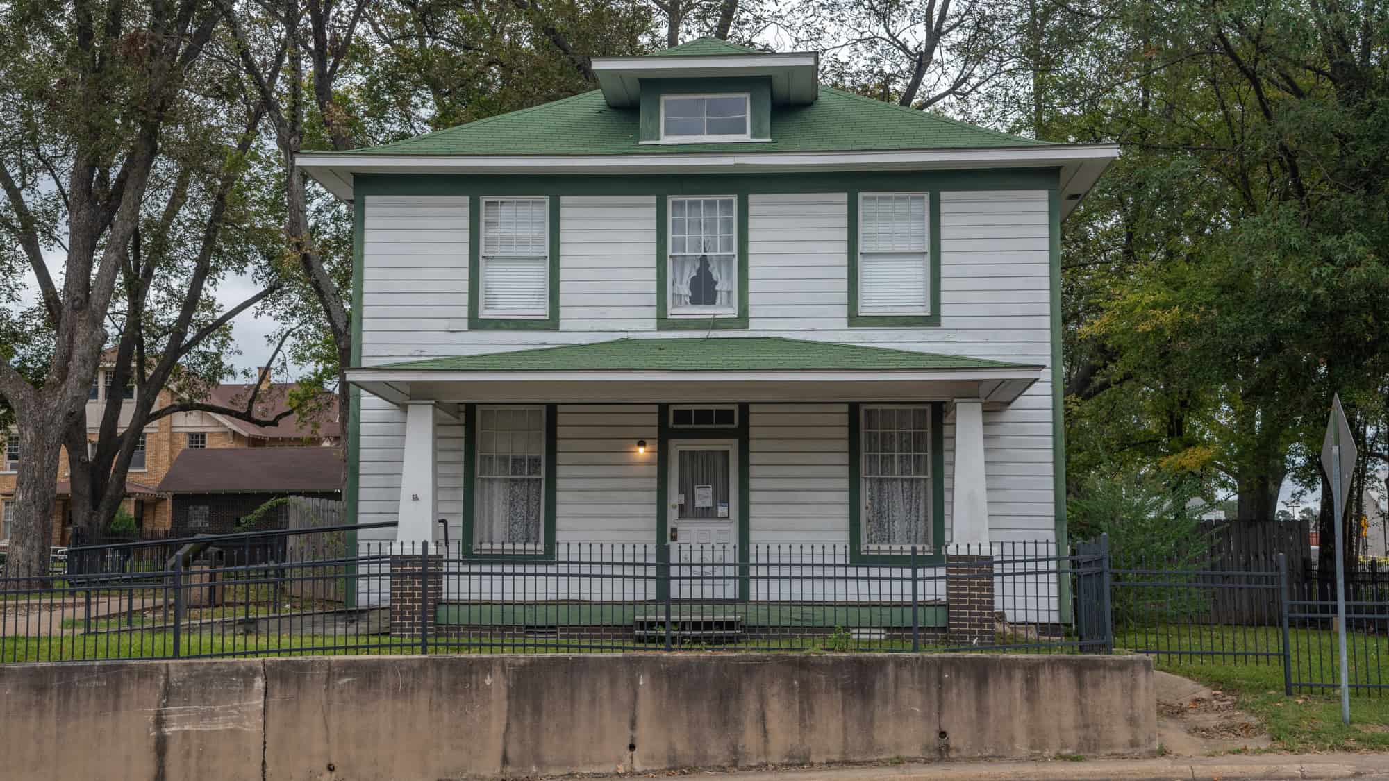 A simple white two-story house with green trim and a green roof stands behind a black iron fence, framed by mature trees in a quiet neighborhood.