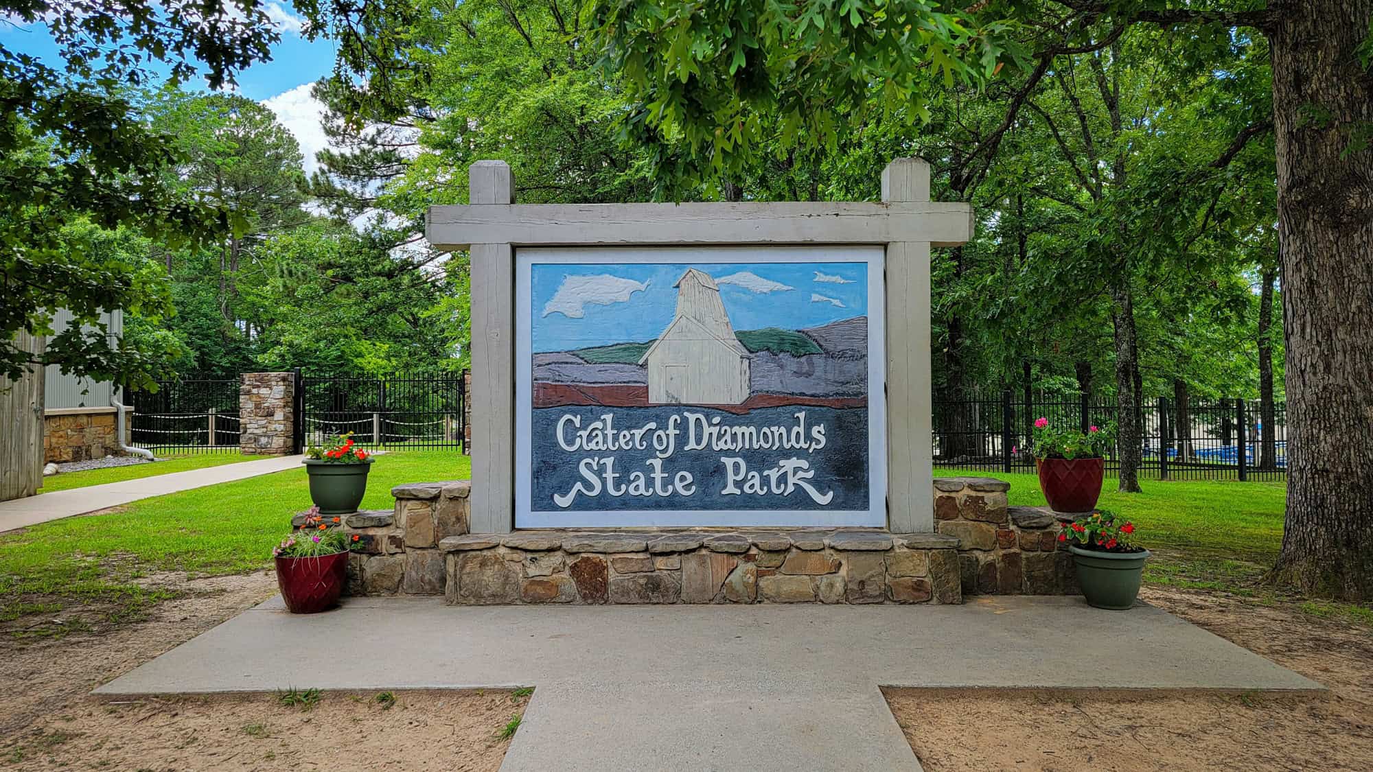 A hand-painted wooden sign for Crater of Diamonds State Park stands among trees and flower pots, welcoming visitors with its rustic charm.