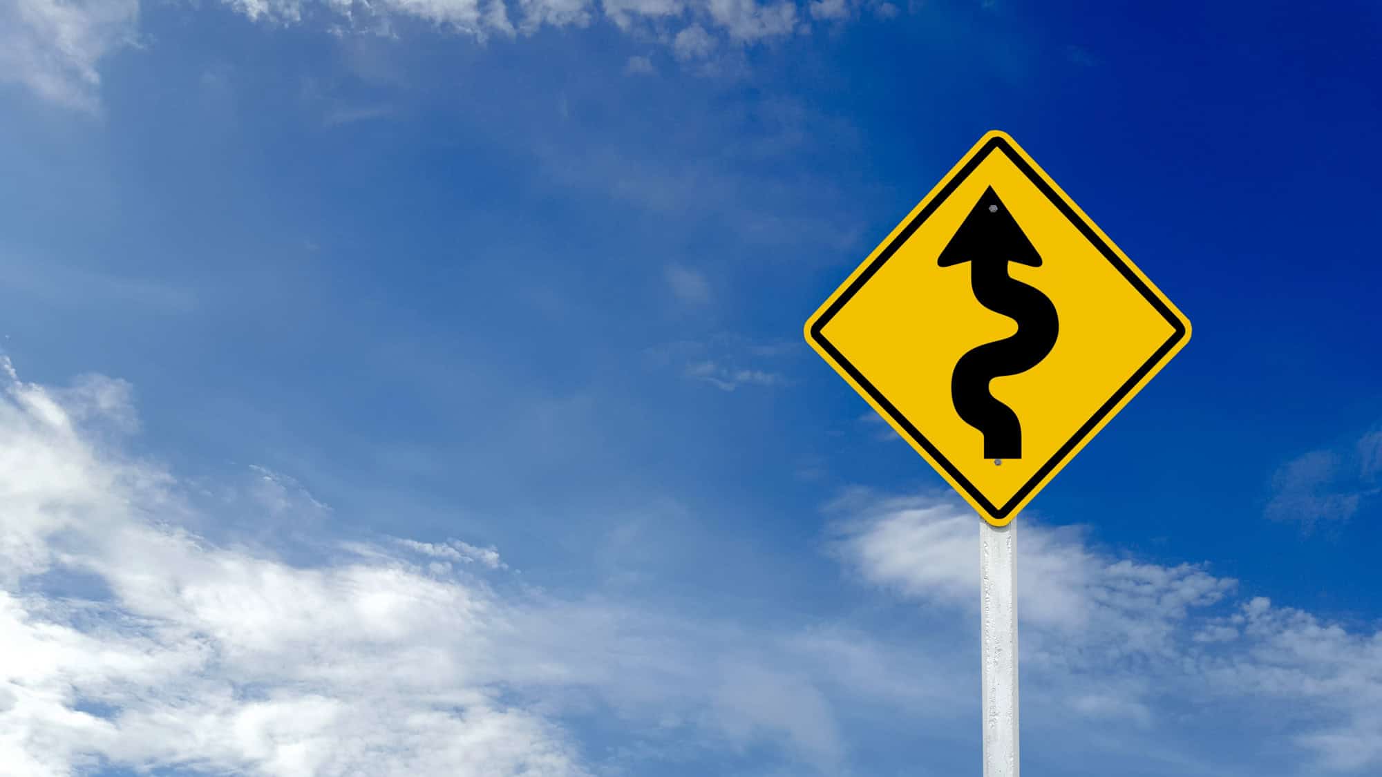 A bright yellow road sign with a black squiggly arrow warns drivers of upcoming winding roads under a clear blue sky.