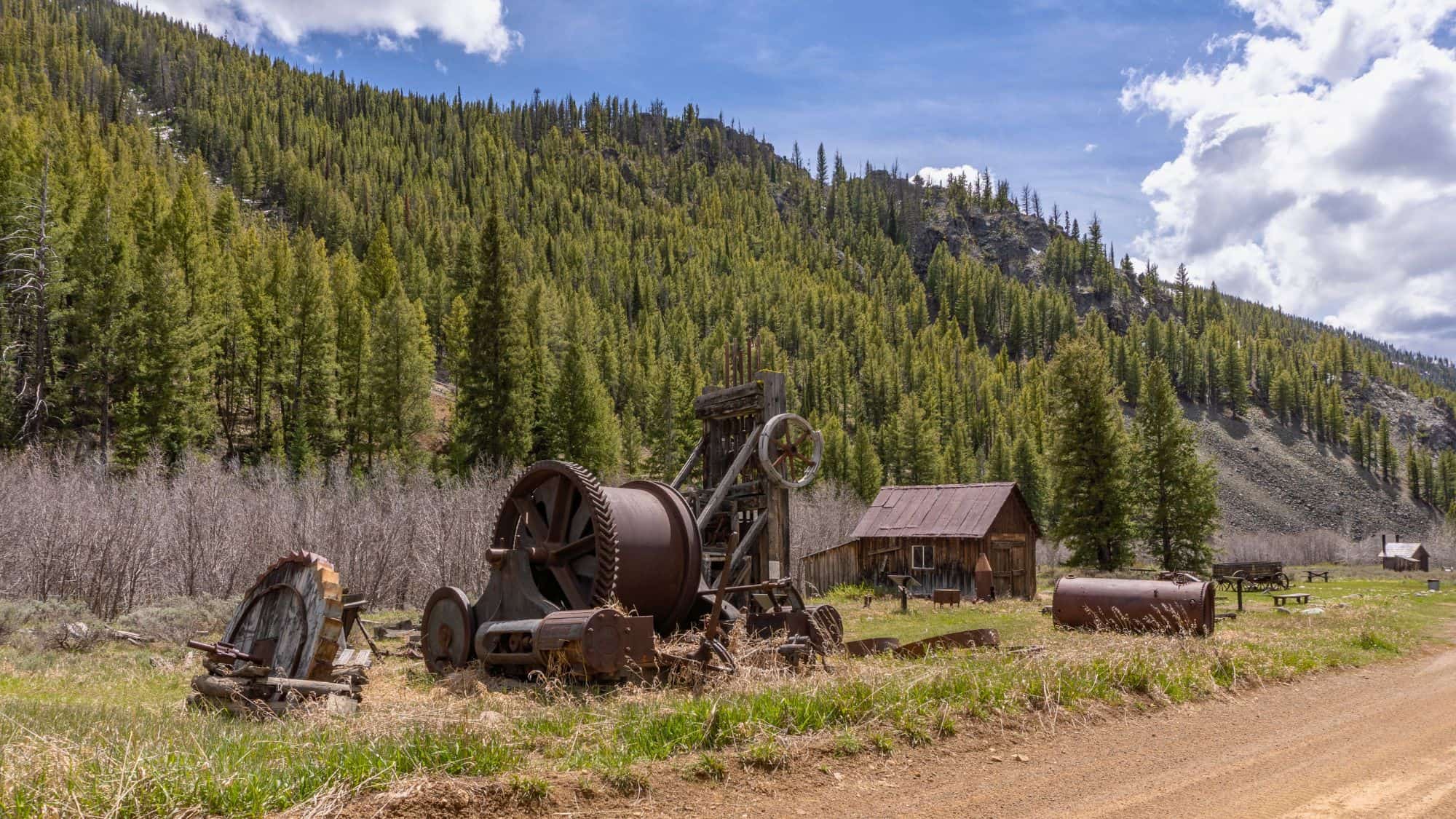 Abandoned mining equipment and rustic cabins sit quietly in a grassy clearing surrounded by pine-covered mountains, evoking the past life of a Montana ghost town.