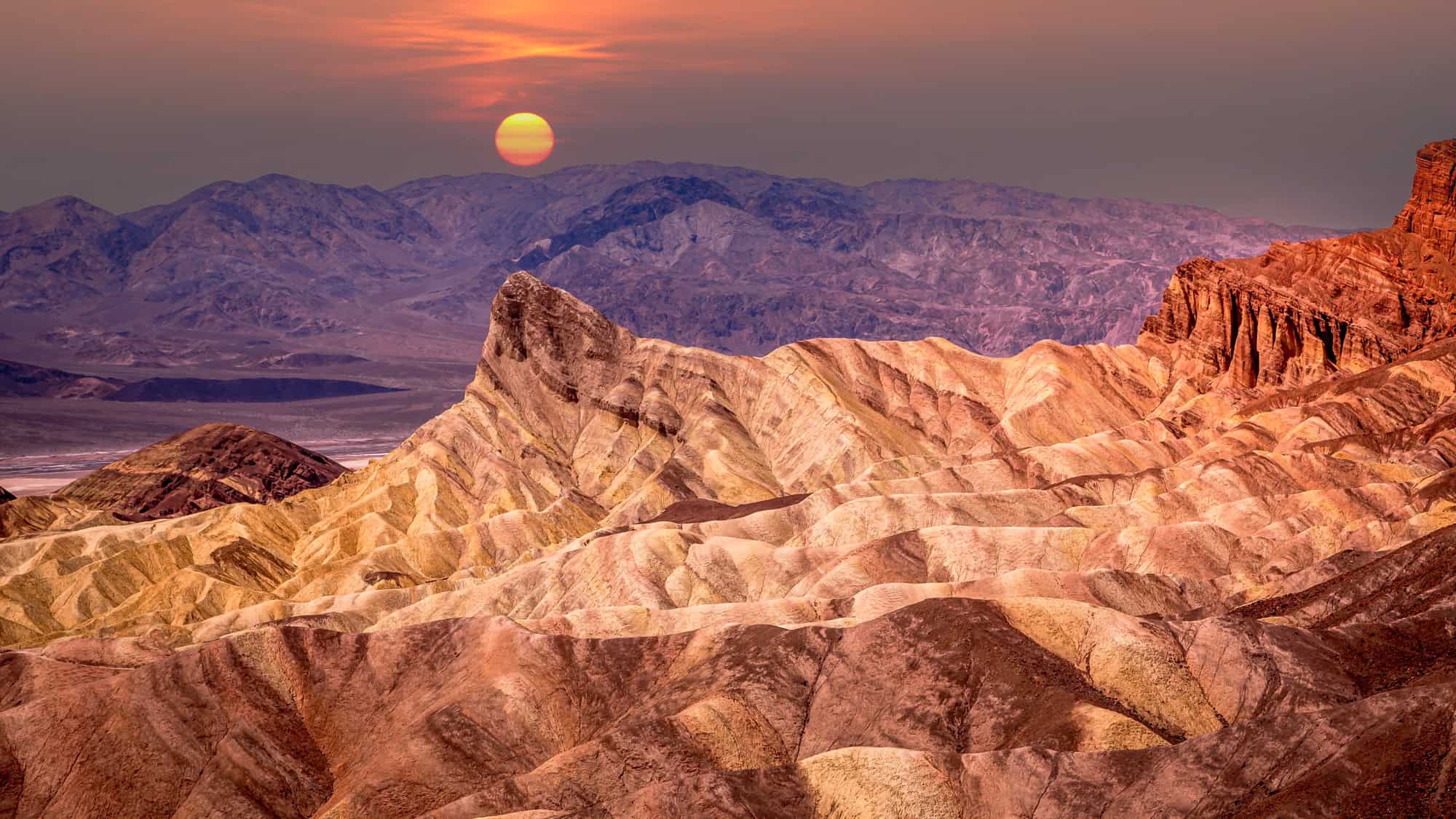 Golden light washes over the rippled badlands of Zabriskie Point in Death Valley as the sun rises above hazy desert mountains in the distance.