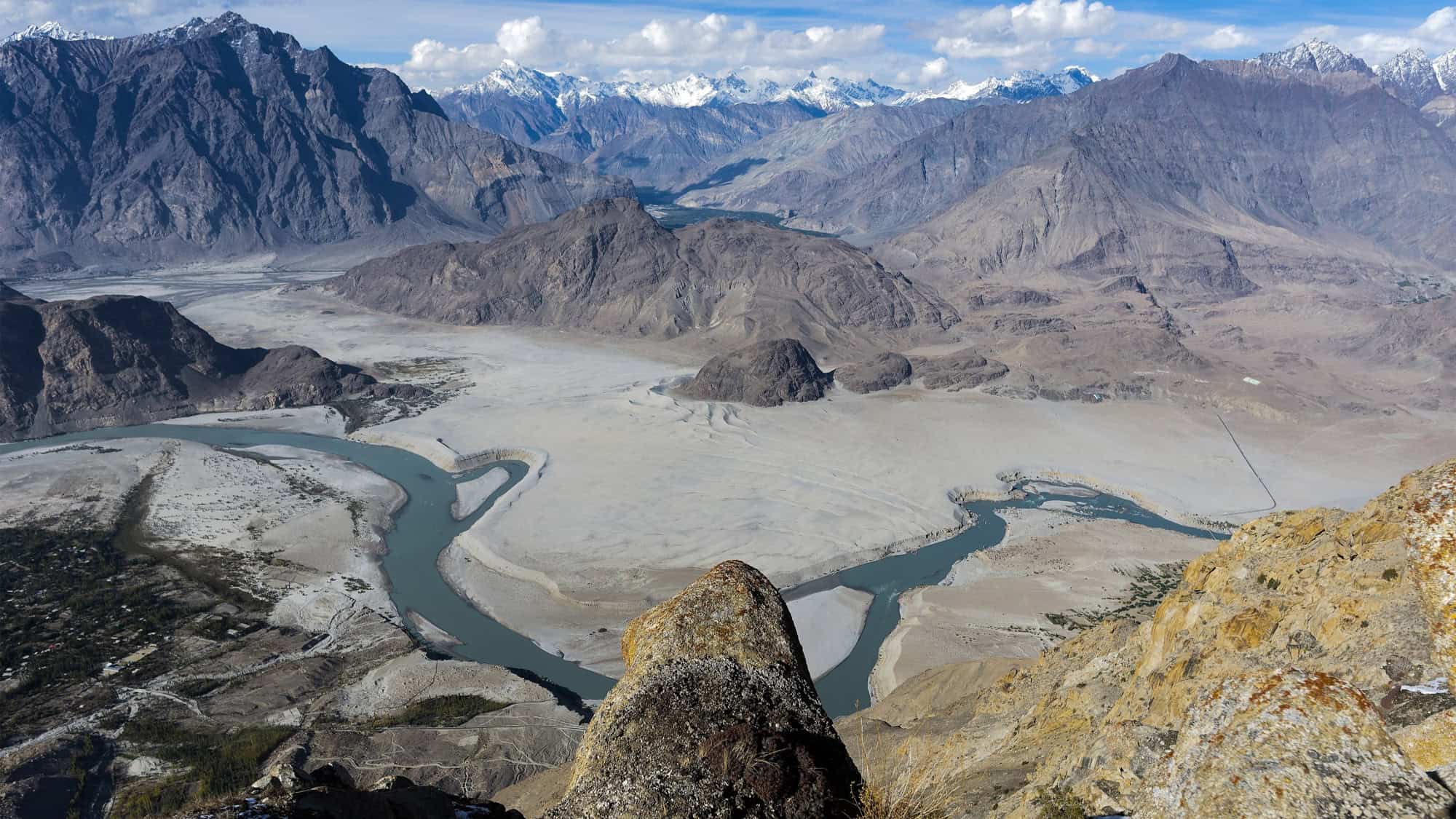 A winding river snakes through a dry, expansive desert valley with steep, rugged mountains and snow-capped peaks under a bright blue sky.