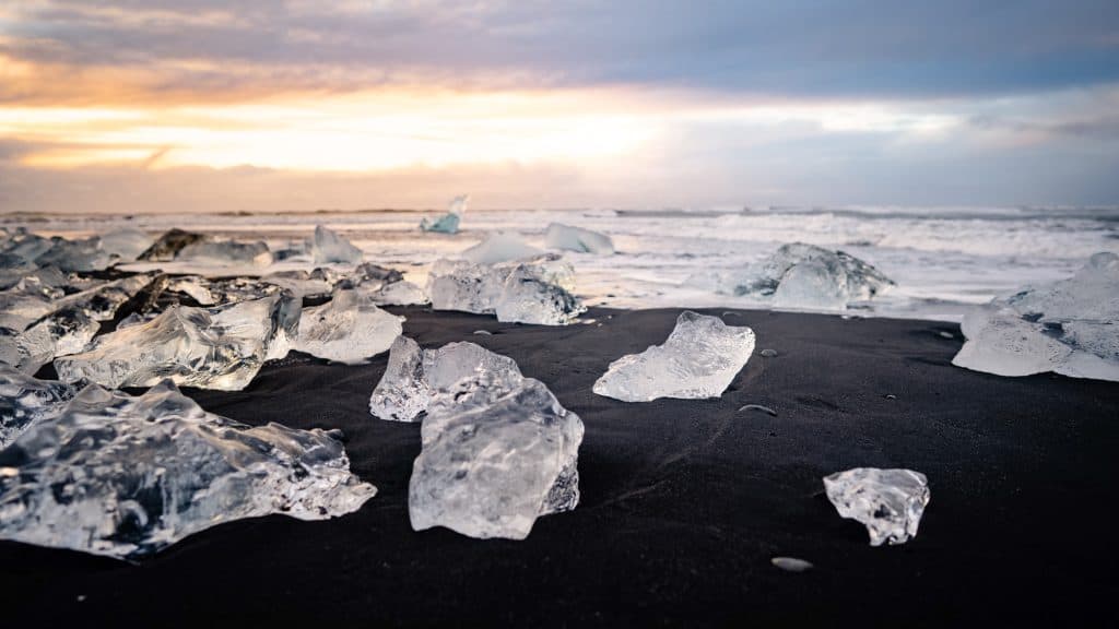Clear ice fragments lie scattered across black volcanic sand with waves crashing in the background under a pastel-colored sky.