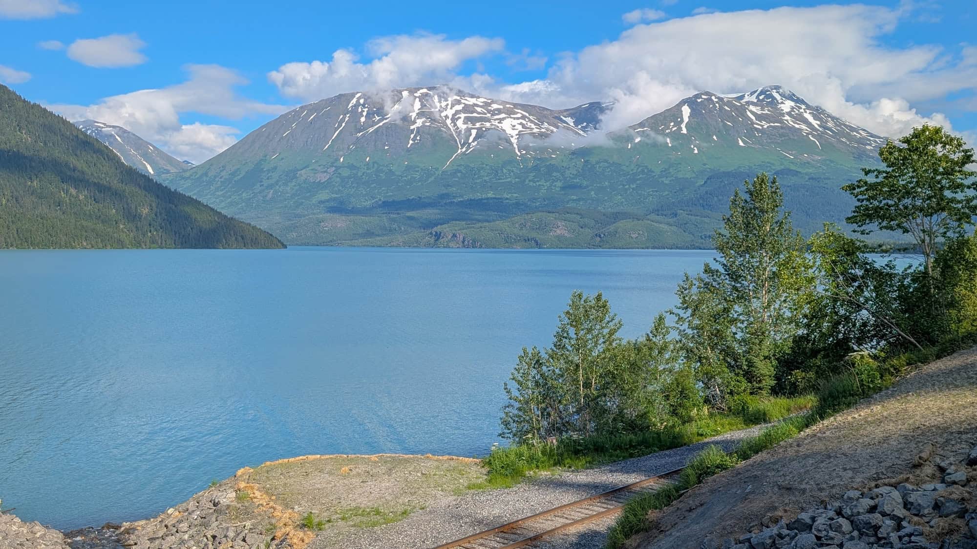 A wide, calm lake reflects a brilliant blue sky and is surrounded by lush green slopes and snow-dusted mountains in the background.