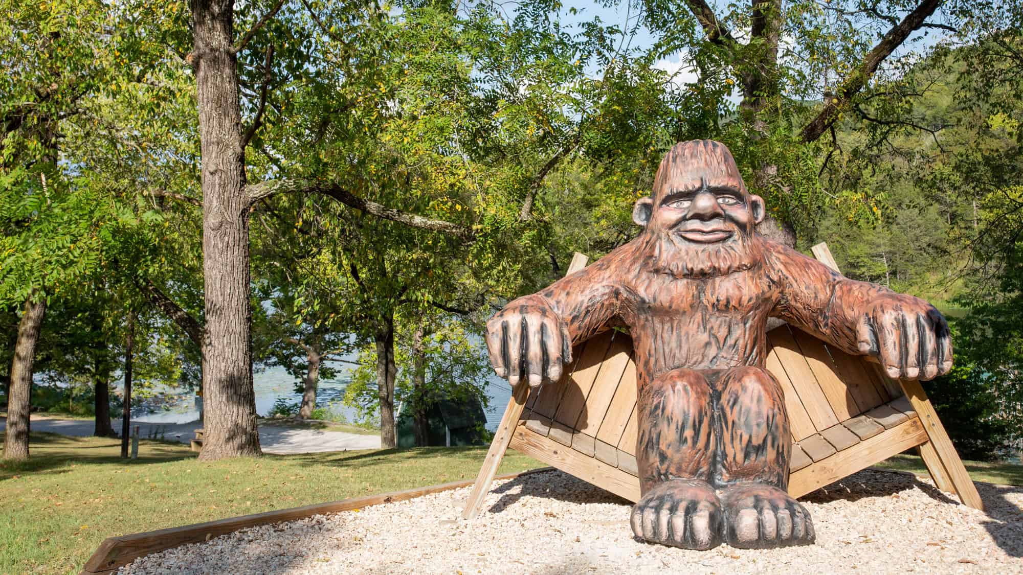 A playful wooden bench shaped like Bigfoot invites visitors to relax under shady trees near a peaceful lakeside setting.