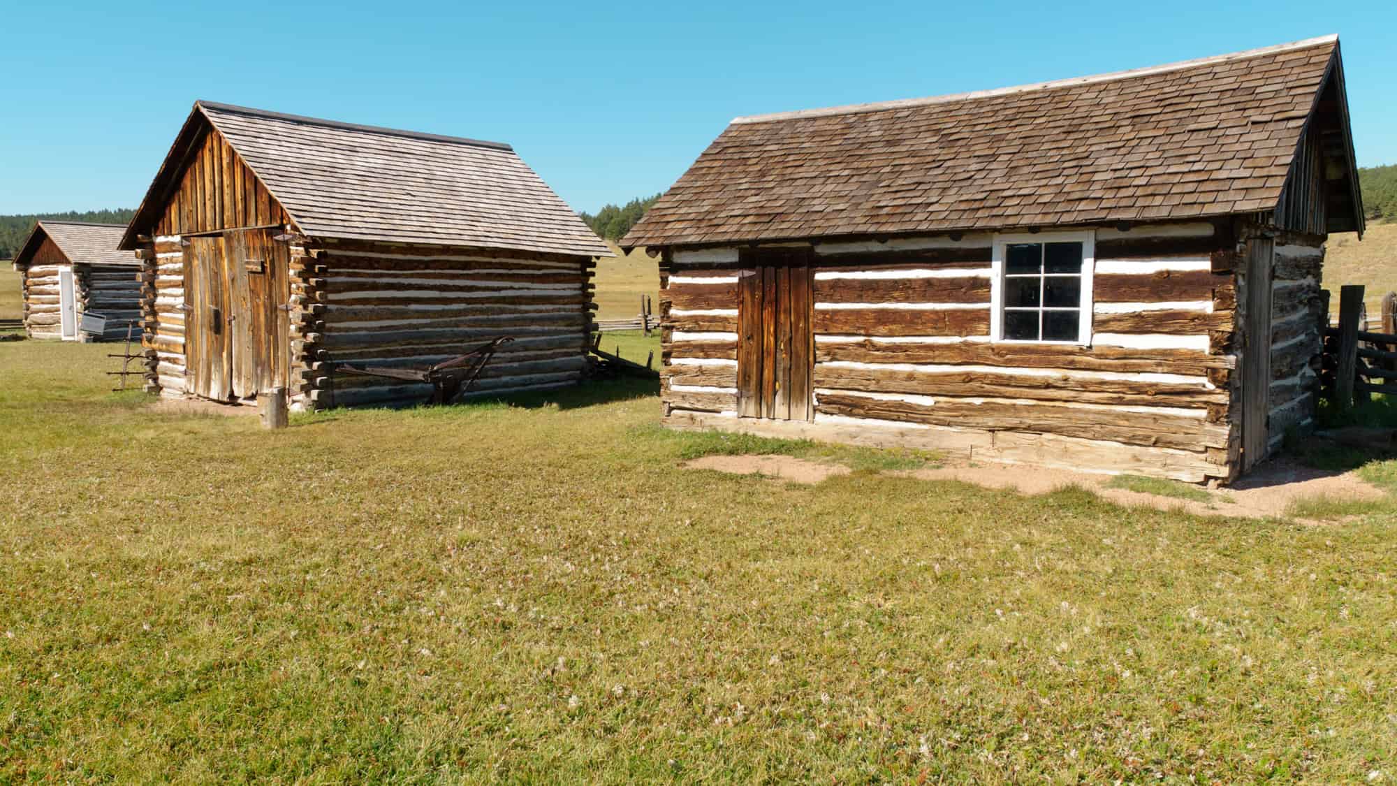 Two rustic log cabins with pitched roofs sit on a grassy plain, part of a preserved pioneer homestead under a bright blue sky.