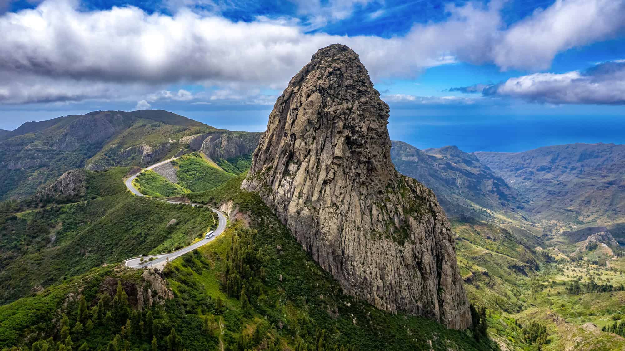 A towering rock spire rises from a green mountain ridge, with a winding road carving through the landscape under scattered clouds and coastal light.