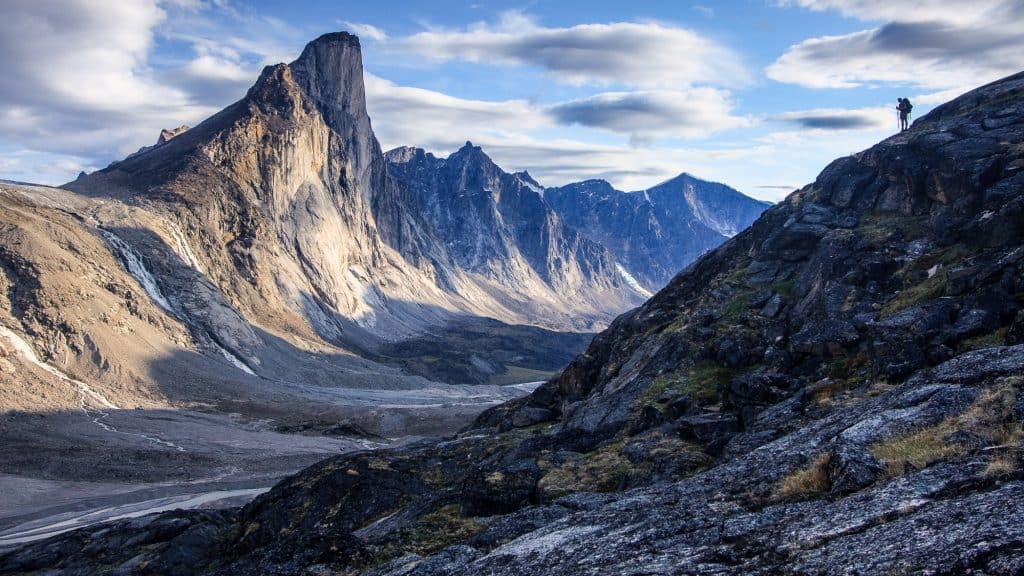 A lone hiker stands atop a rocky slope, gazing at the towering, dramatic peaks of a mountain range with sheer cliffs and scattered snow patches.