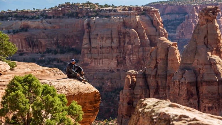 A hiker in dark clothing sits at the edge of a red rock cliff, taking in the breathtaking, sheer-walled canyons of Colorado National Monument.