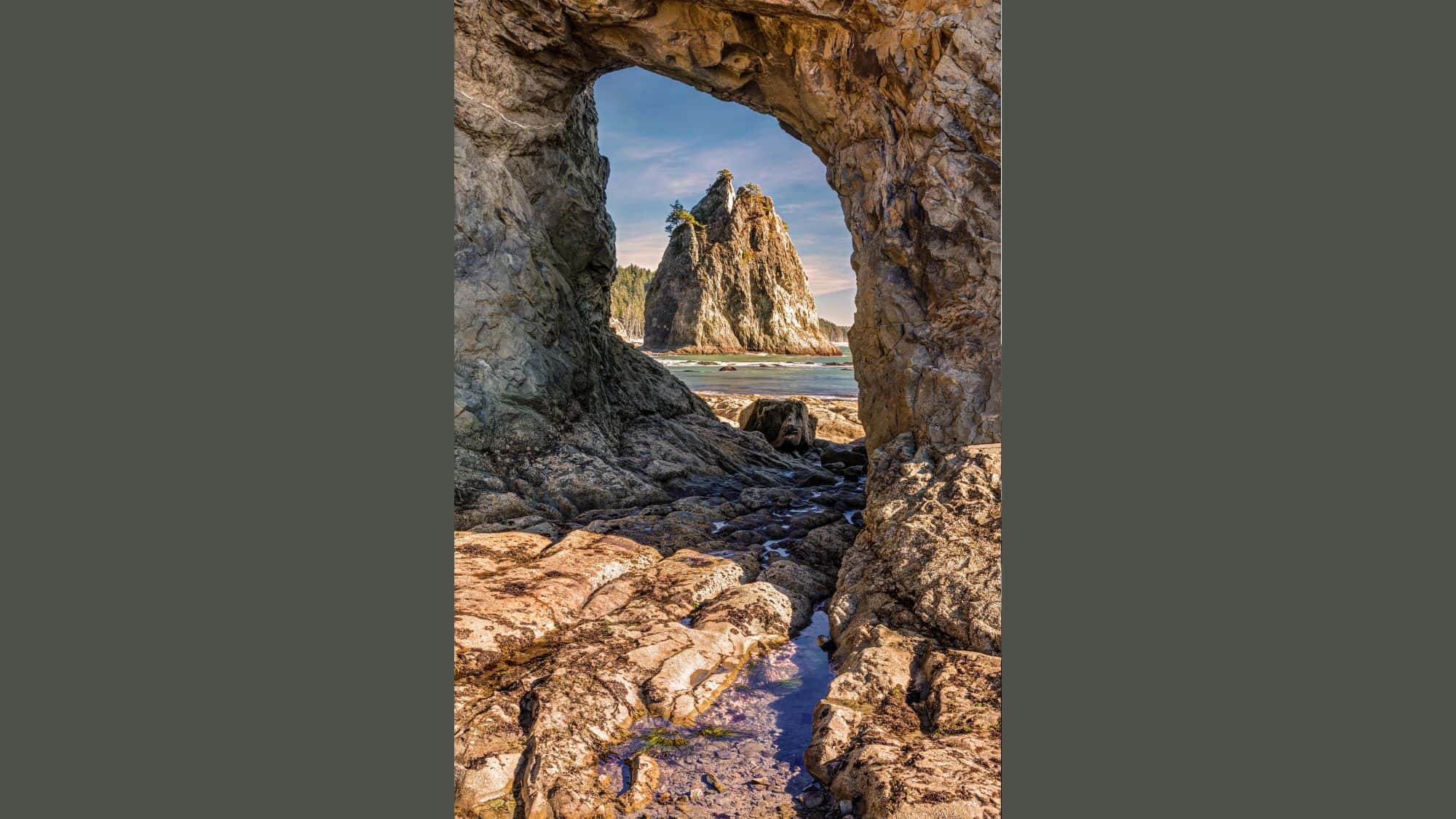 A towering sea stack is framed by a rocky arch in the foreground, with tide pools and rugged textures leading the eye to the beach beyond.