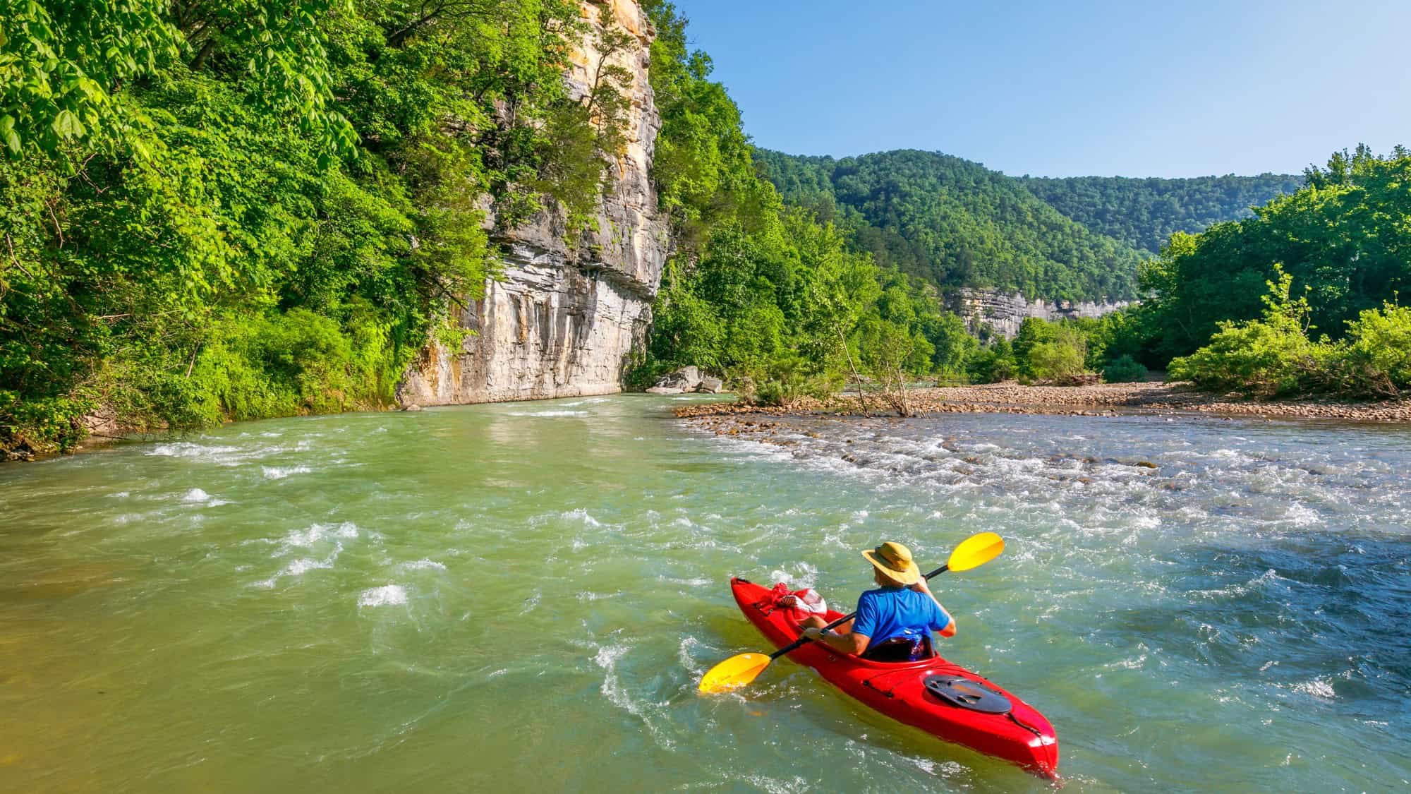 A kayaker paddles through the turquoise waters of the Buffalo National River, surrounded by steep limestone cliffs and dense greenery on a sunny day.