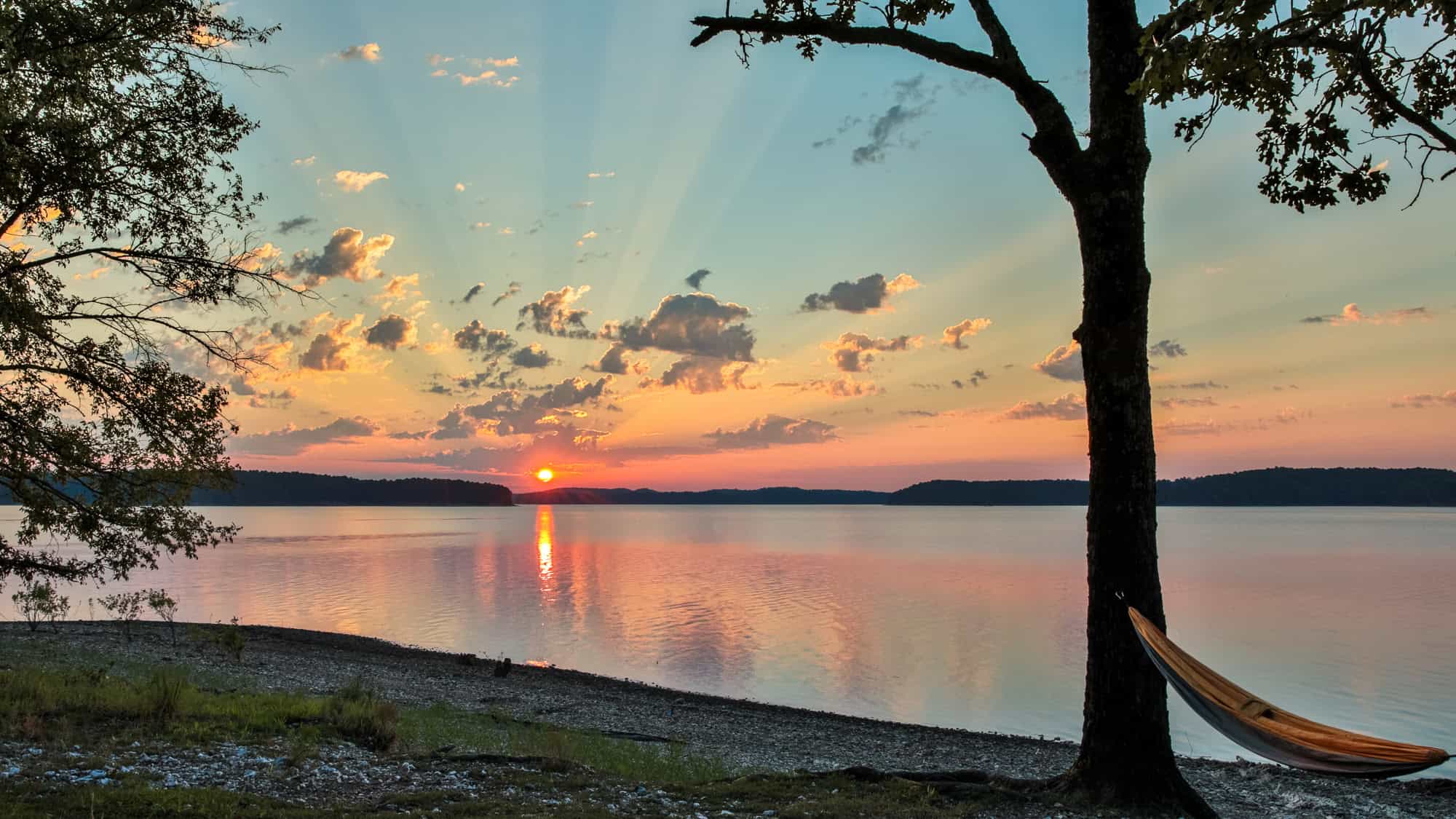 A serene lake reflects the golden glow of a setting sun, with sunrays streaking through scattered clouds and a hammock tied to a tree in the foreground.
